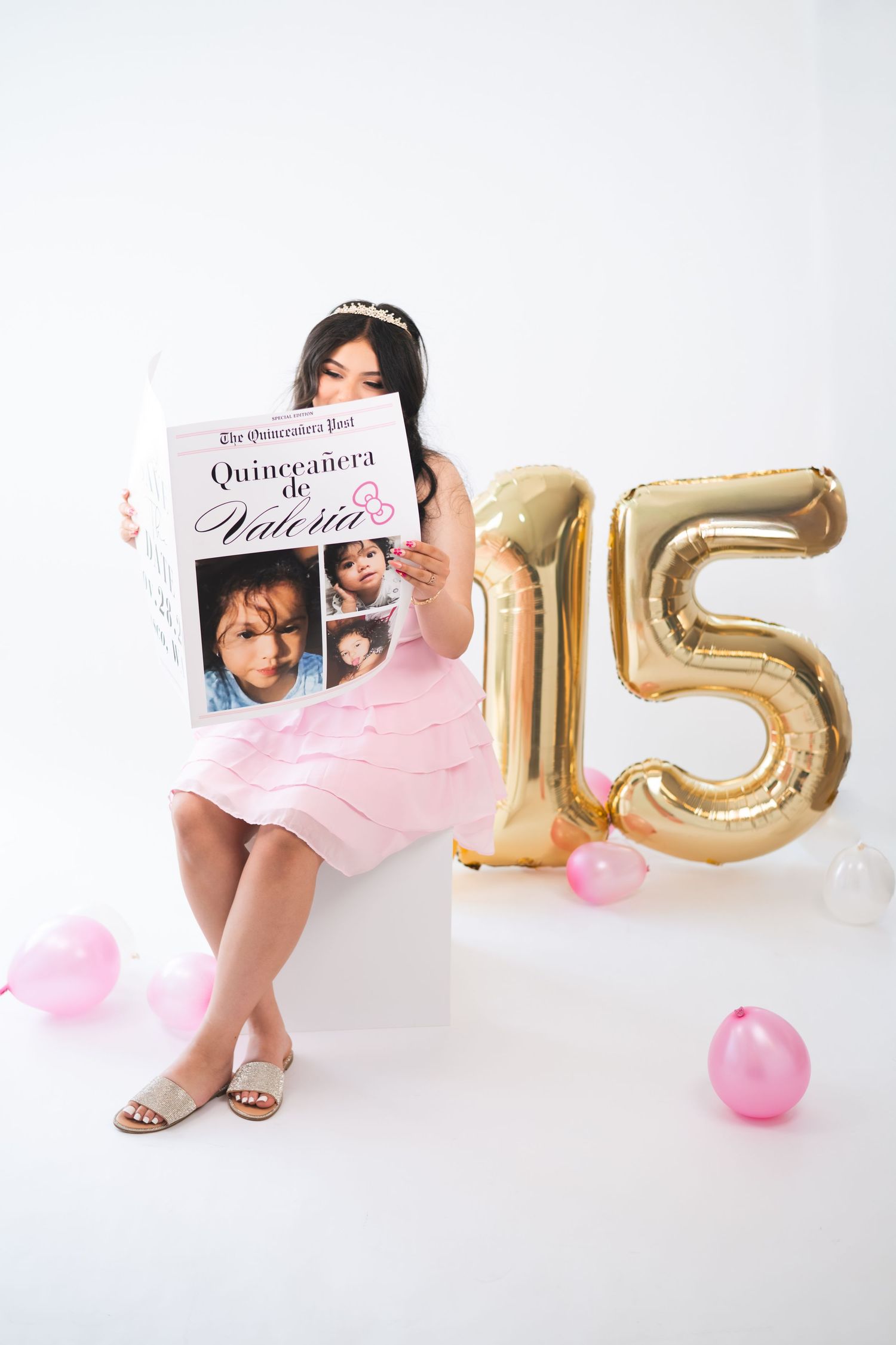 Girl in pink dress holds Quinceañera newspaper beside gold number 15 balloons at a birthday celebration.