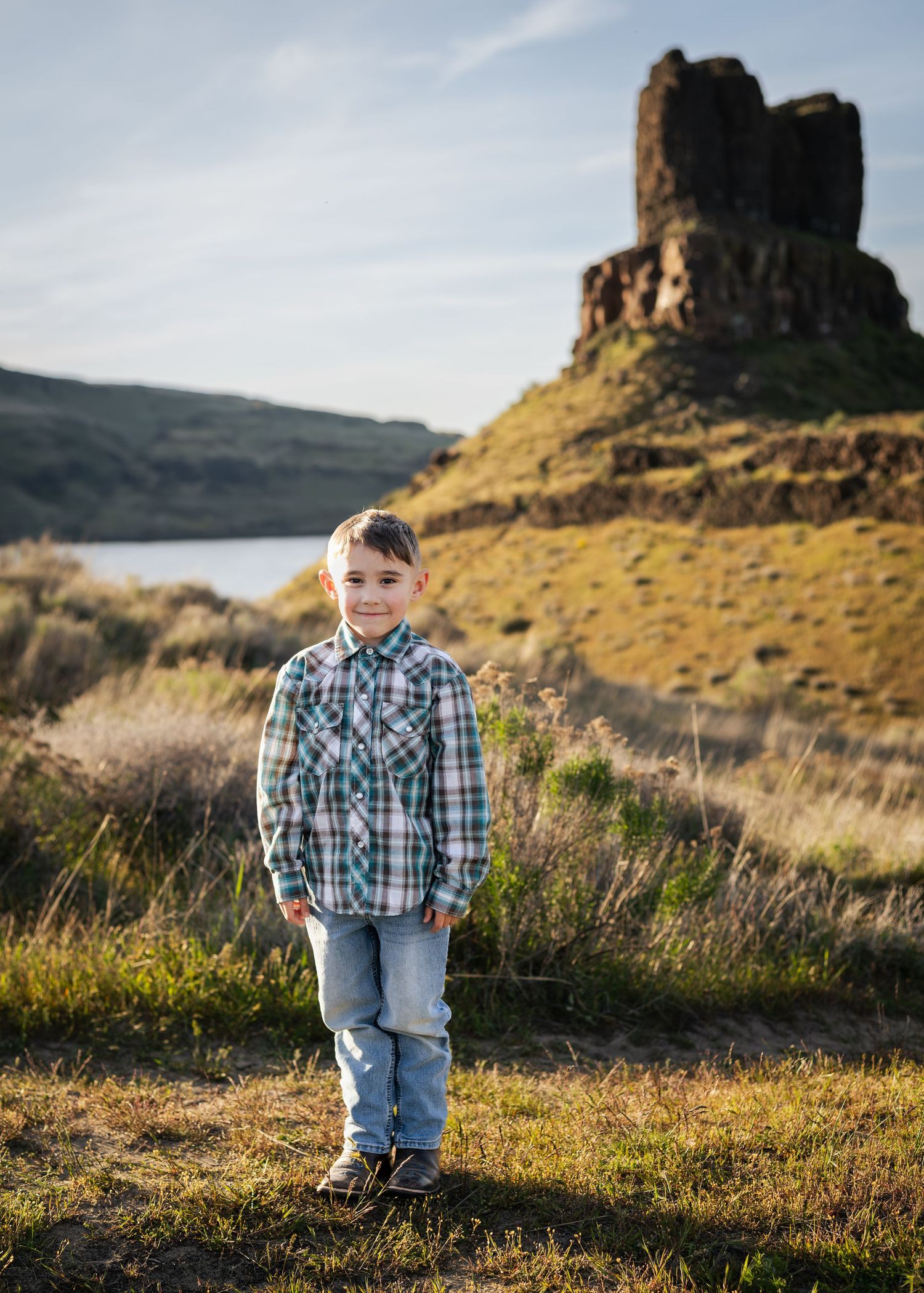 Young boy in plaid shirt and jeans stands on grassy terrain with rocky butte and river in background.