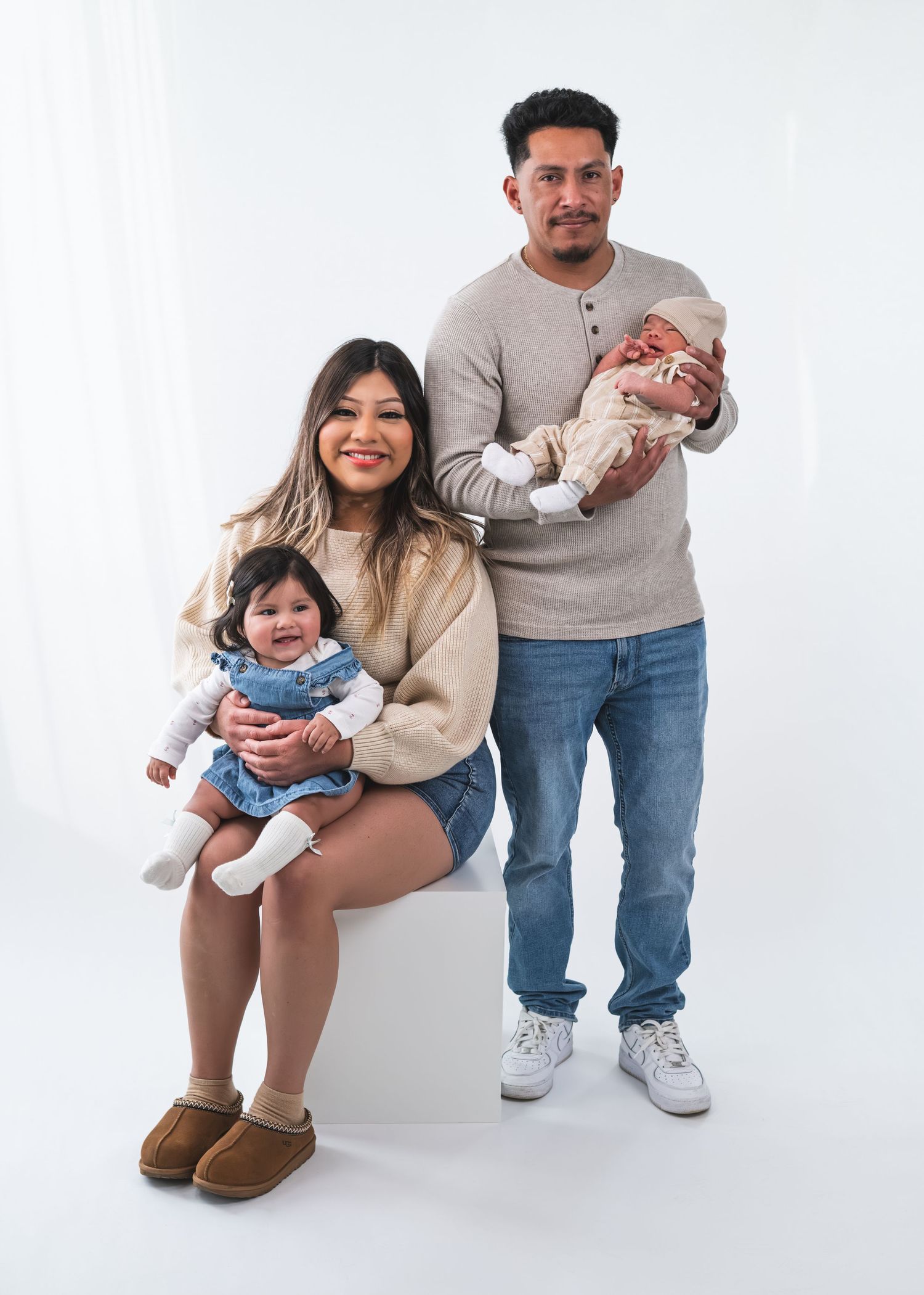 Young family portrait with parents holding a toddler and newborn baby, posing against a white studio background.