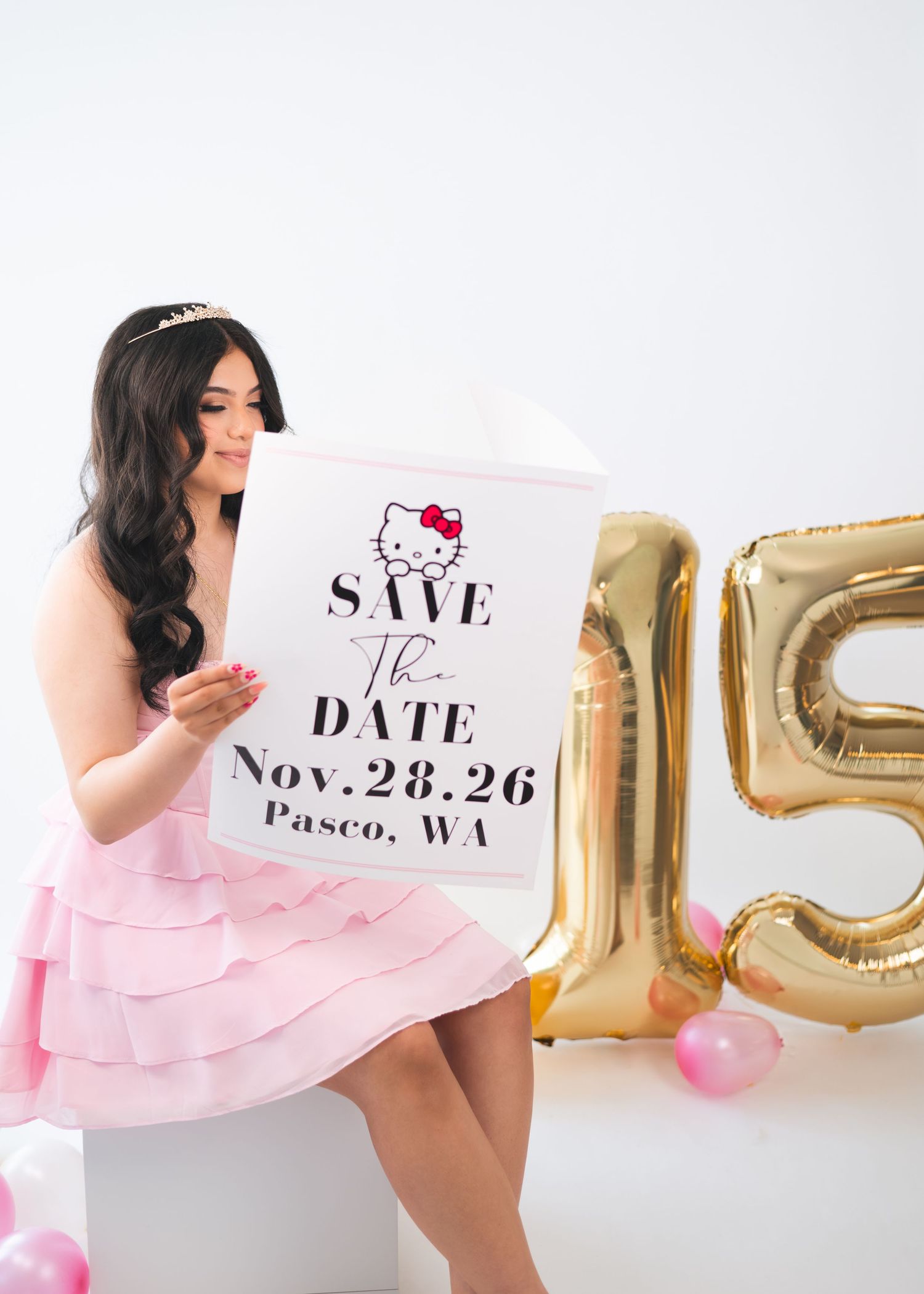 Girl in pink ruffle dress holds Hello Kitty Save the Date sign beside gold number 15 balloons for Quinceañera.
