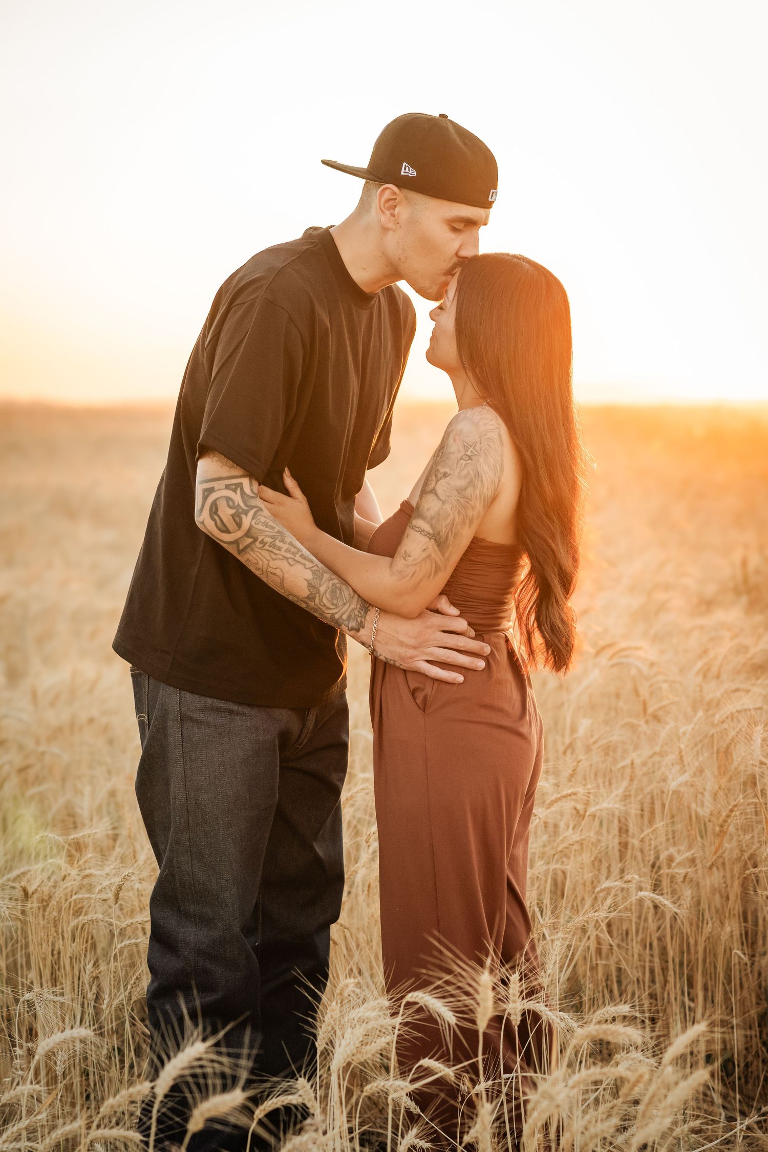 Couple sharing a romantic kiss at golden sunset in a wheat field, man with tattoos in black tee and cap, woman in brown dress.