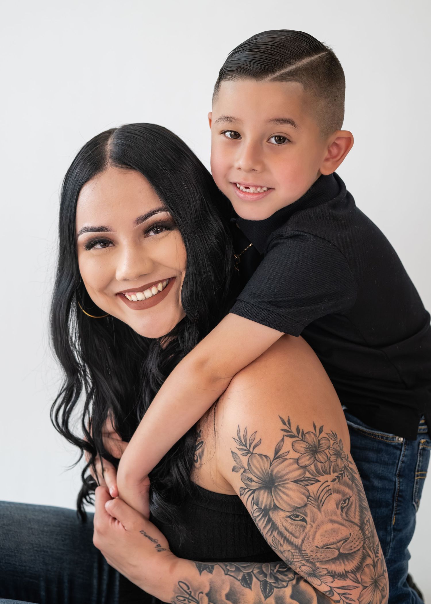 A young woman with long black hair and tattoos smiles while a boy in a black shirt hugs her from behind.