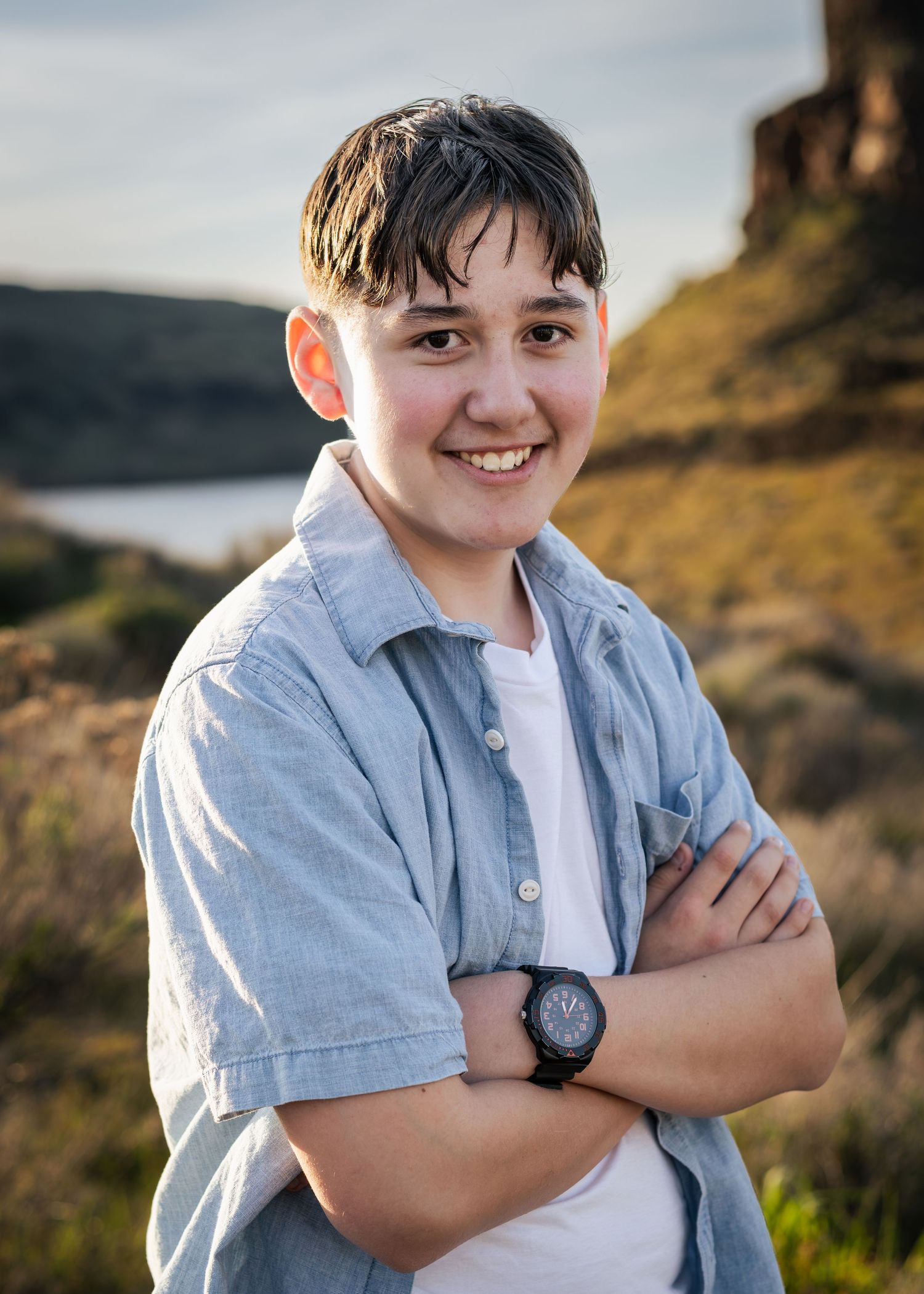 Smiling teenage boy with crossed arms wearing a blue shirt and watch outdoors near a rocky hillside.