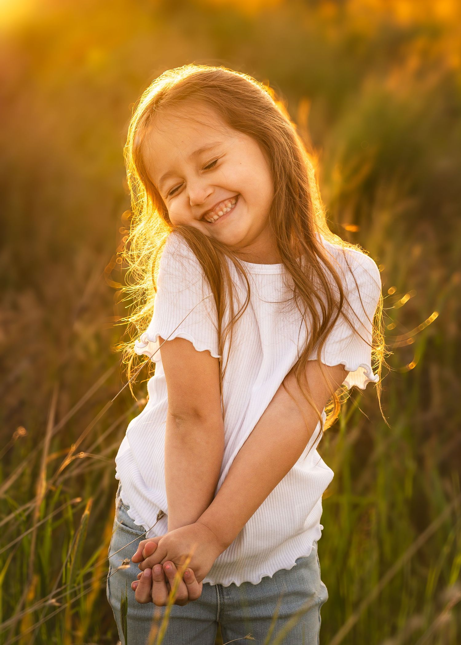 A young girl in a white top and jeans laughs joyfully in a sunlit field during golden hour.