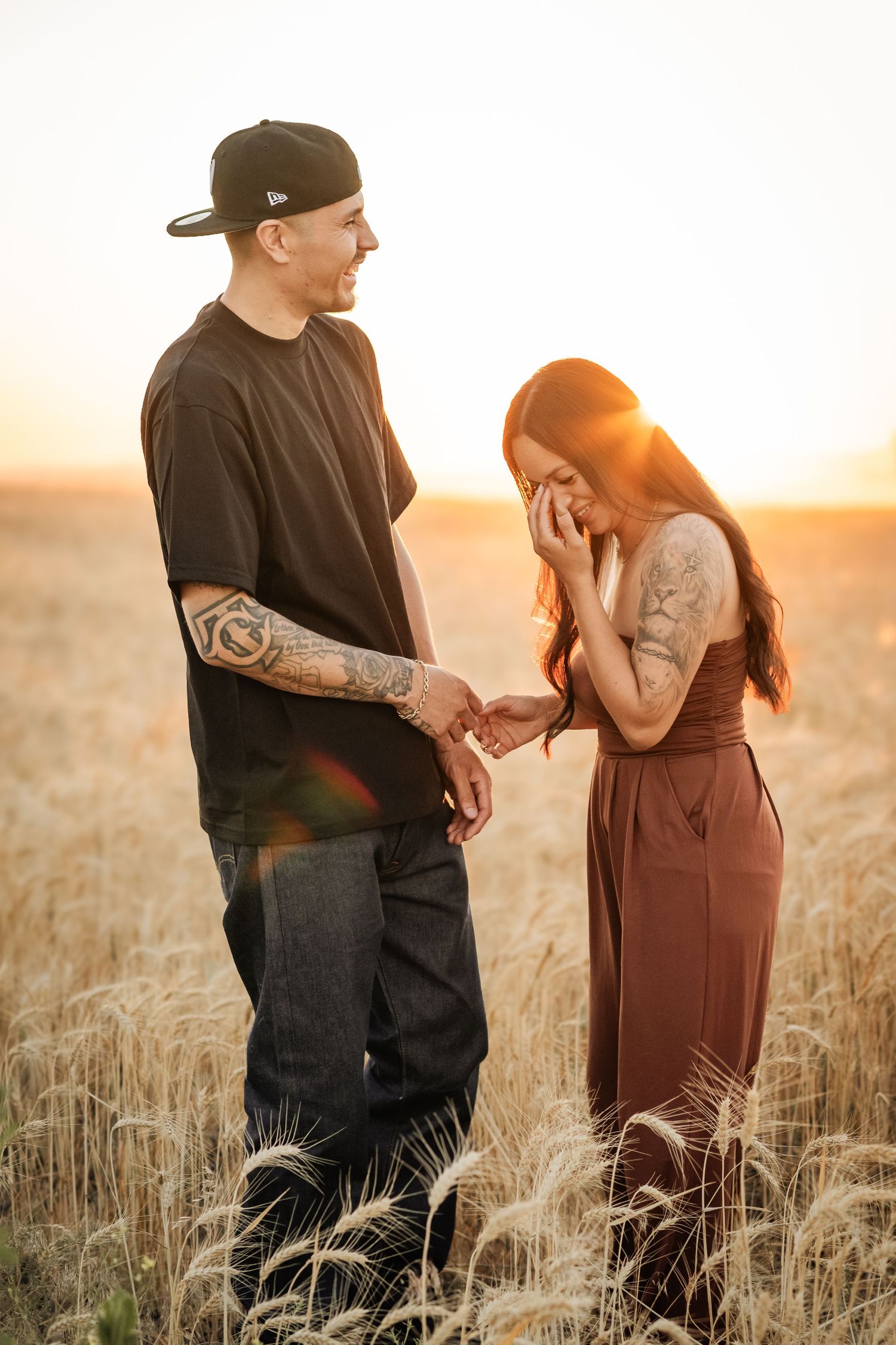 Couple holding hands at golden sunset in wheat field, man in black tee and cap, woman in brown dress with tattoos.