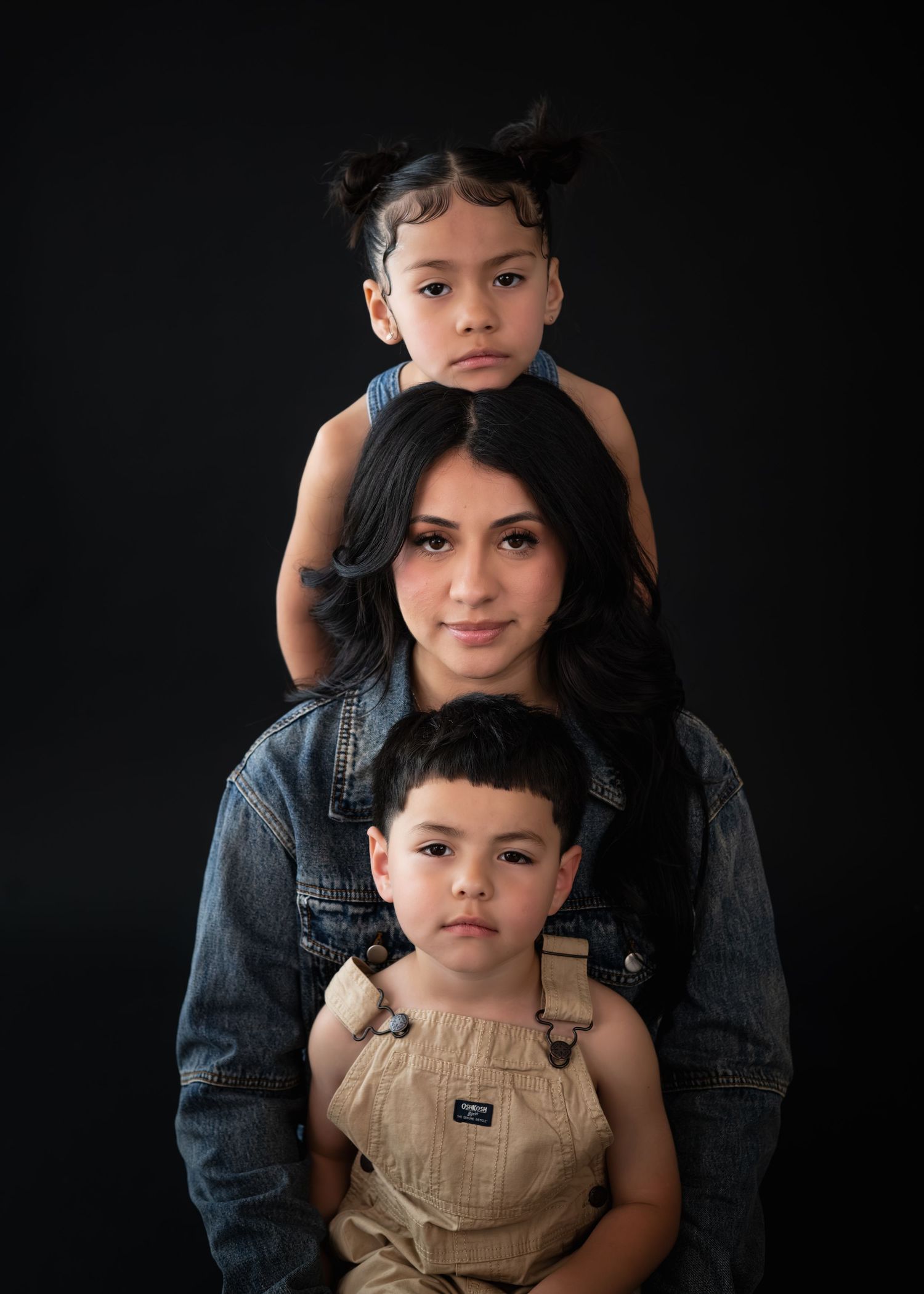 A mother poses with two young children stacked vertically against a dark background in a studio portrait.