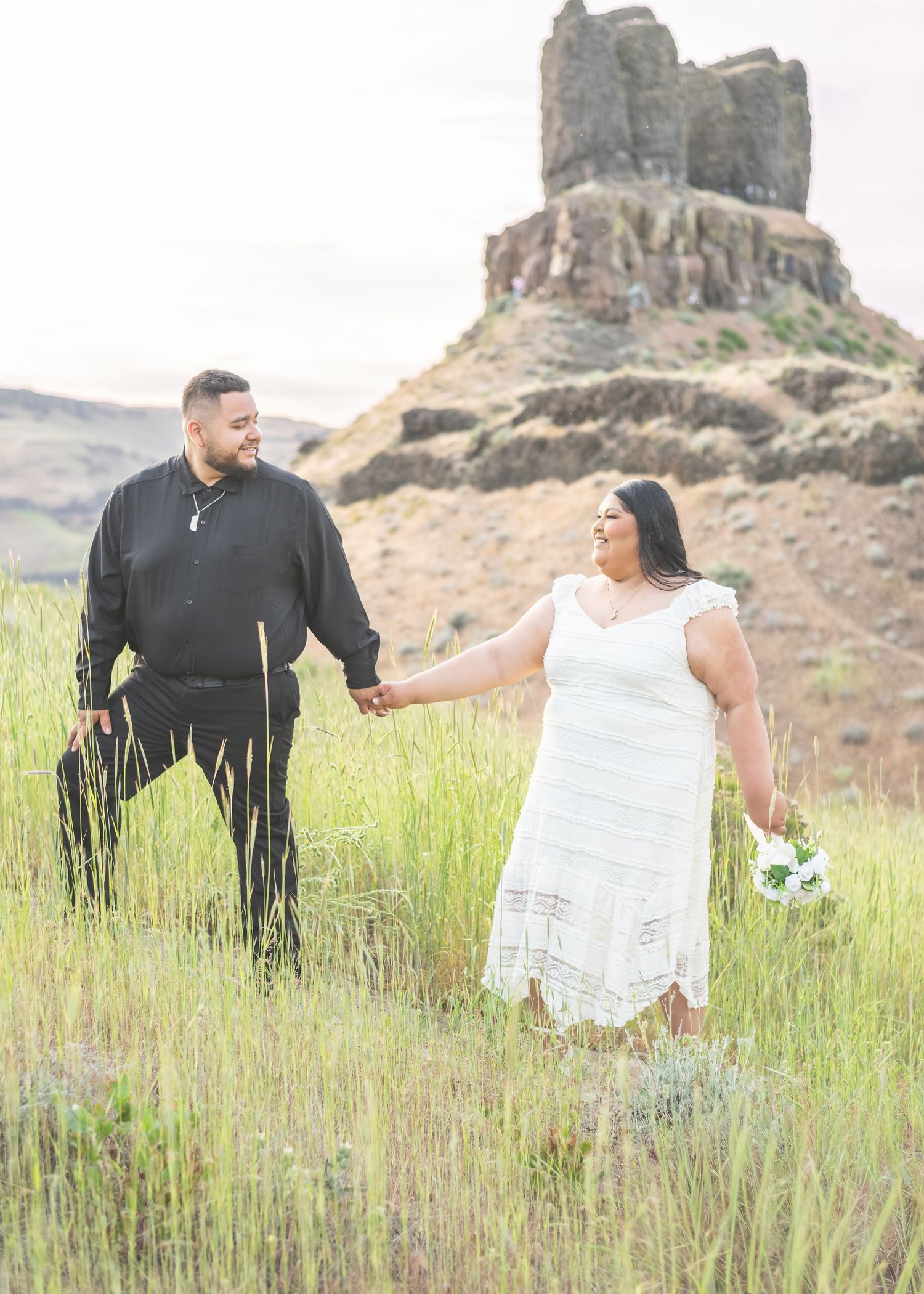 Couple holding hands in a field with a rocky butte backdrop, bride in white dress with bouquet, groom in black attire.