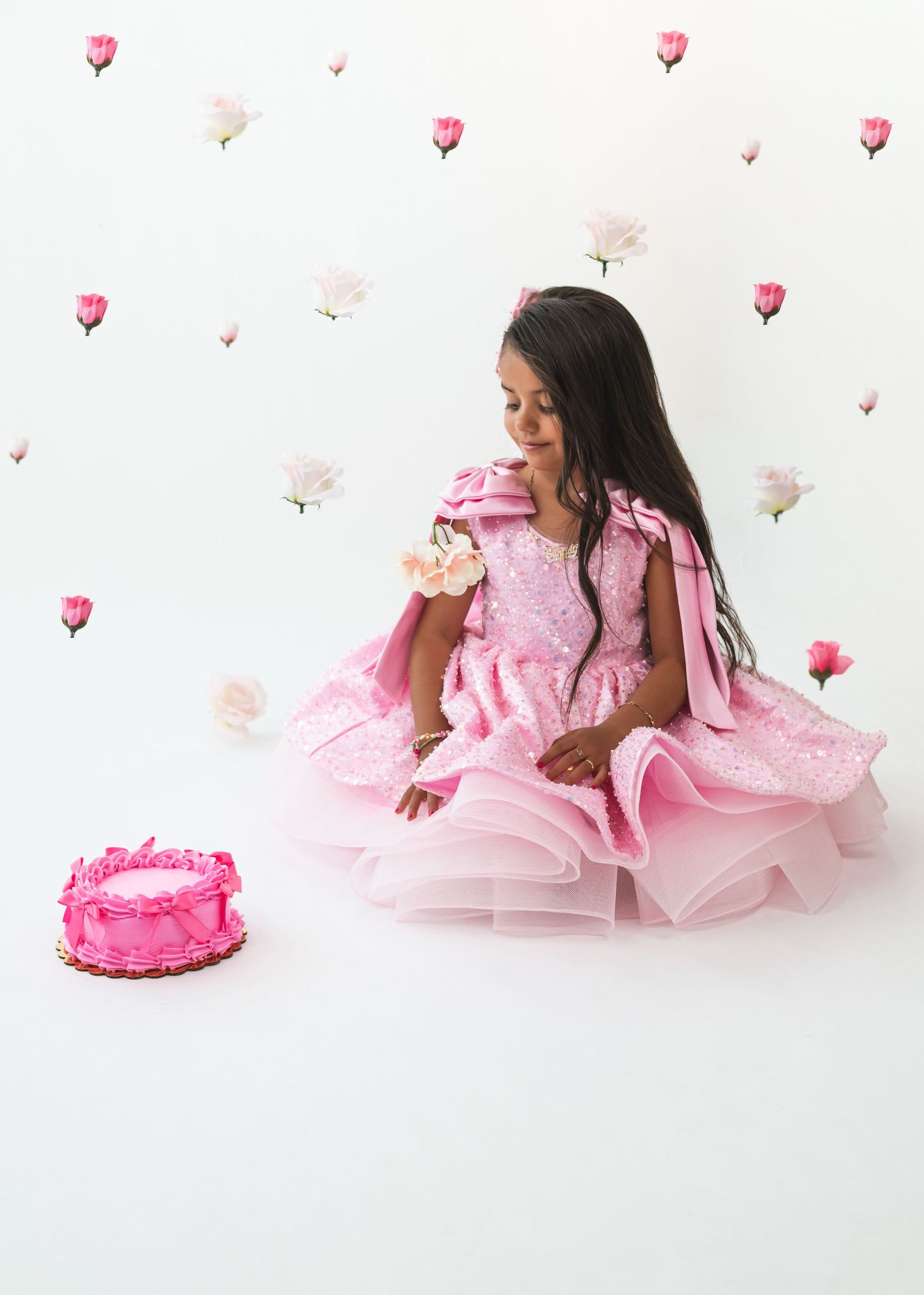 A young girl in a pink tulle dress sits beside a pink birthday cake against a white wall with rose decorations.