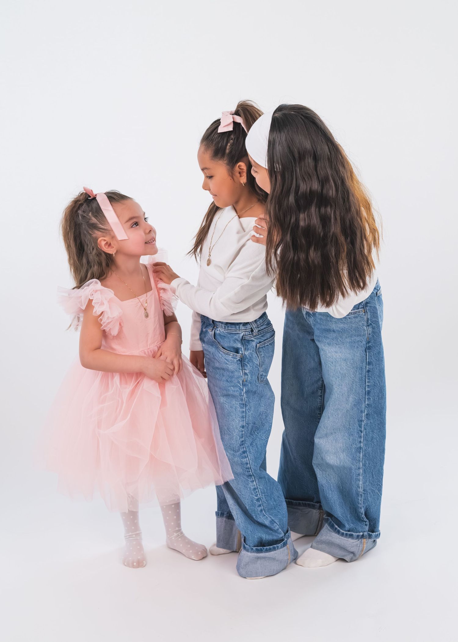 Three young girls in pink and denim outfits pose together on a white background, wearing pink hair accessories.