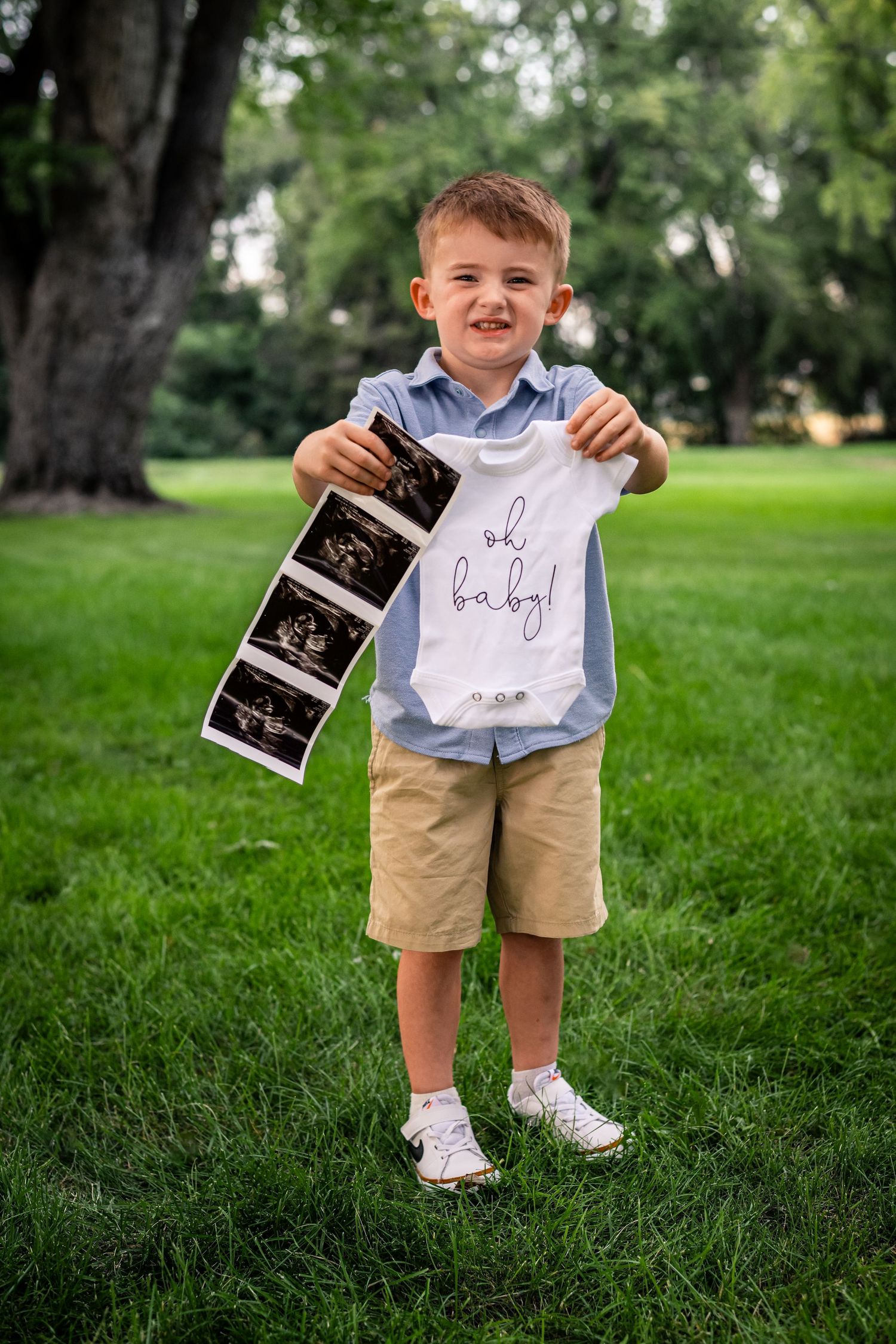 Child in khaki shorts holds ultrasound photos and pregnancy announcement sign in a park setting.