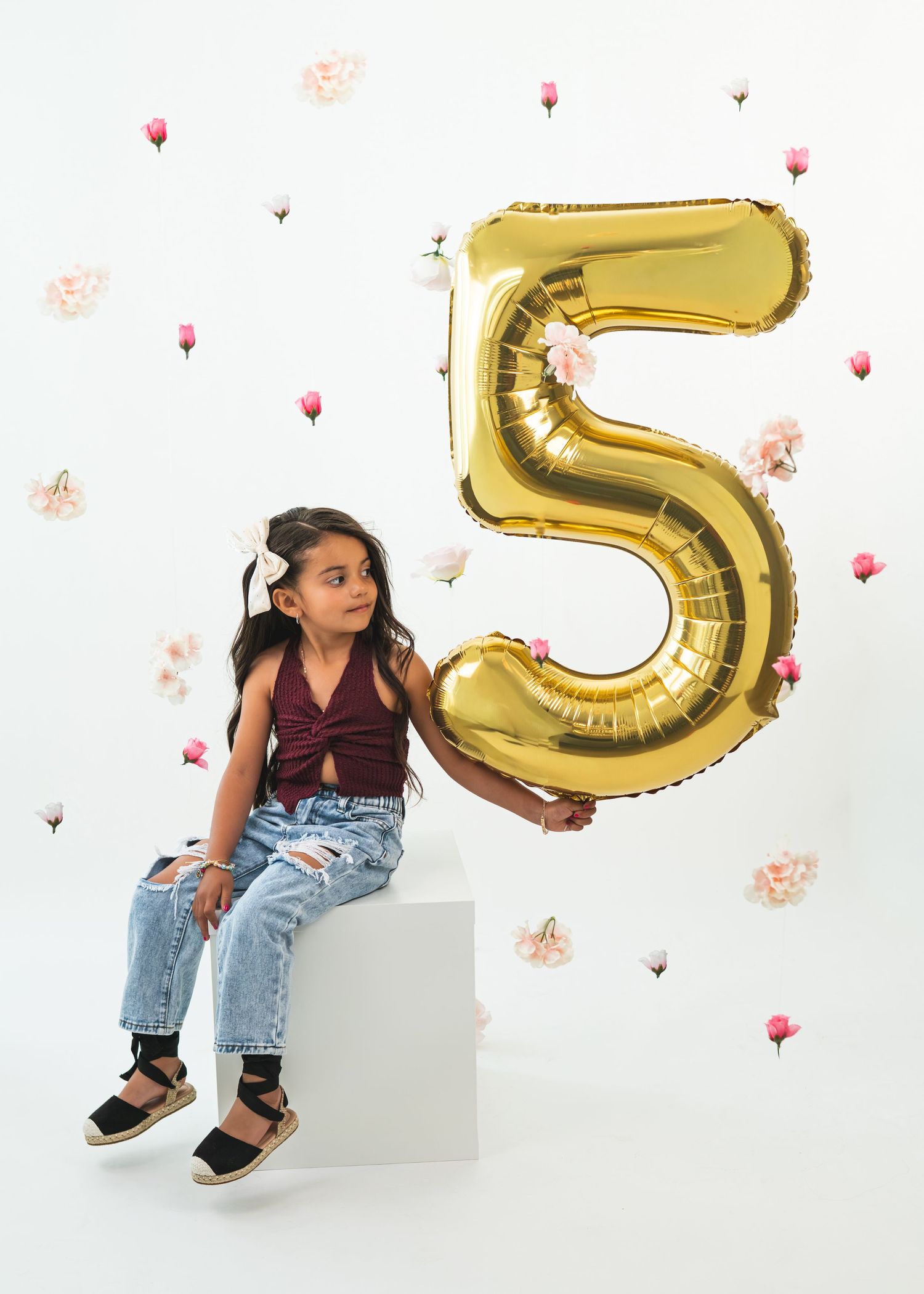 A young girl sits on a white cube holding a large gold number 5 balloon, wearing jeans and a maroon top.