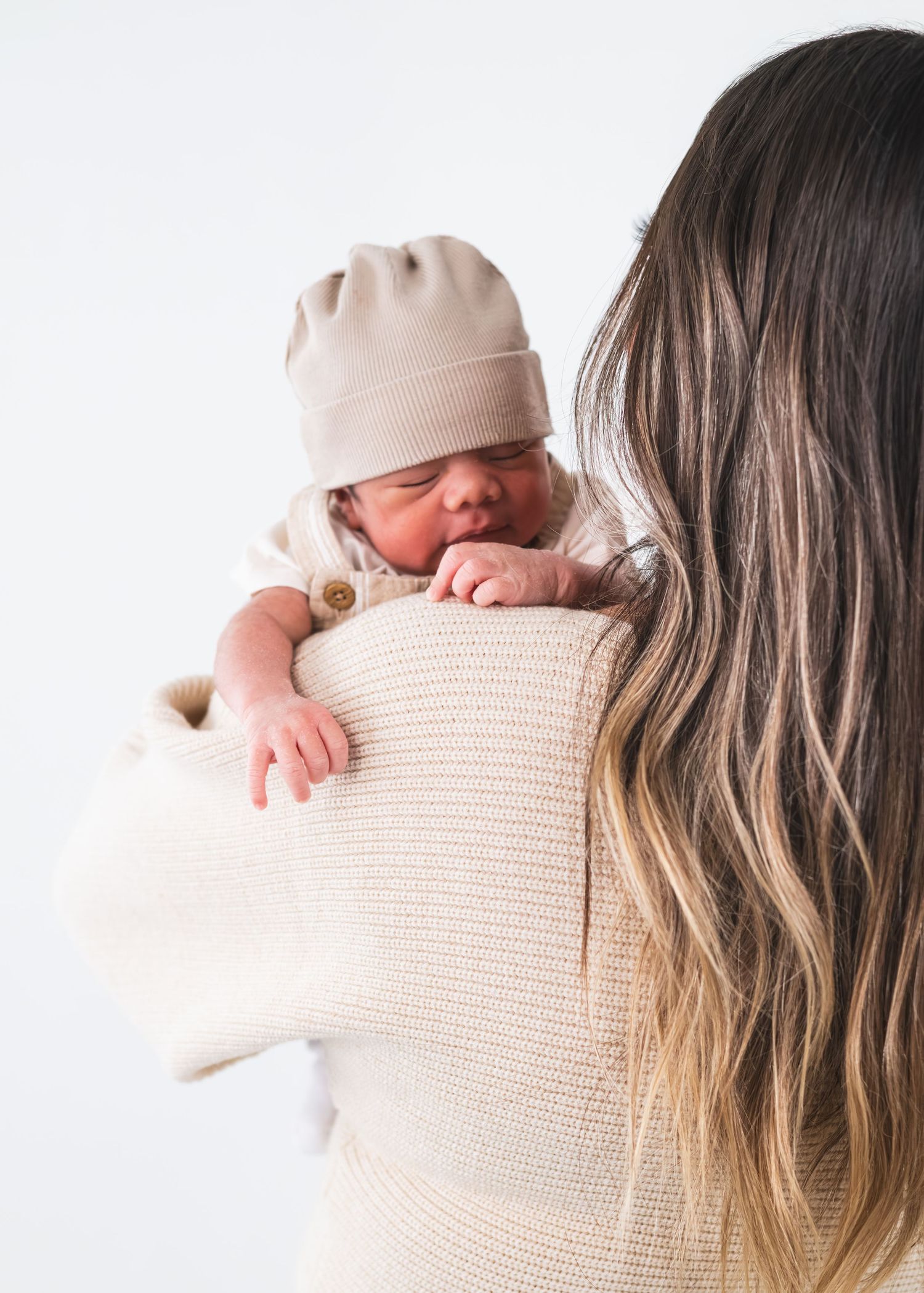 A mother with long ombre hair holds a sleeping newborn baby in a beige knit outfit and hat against a white background.