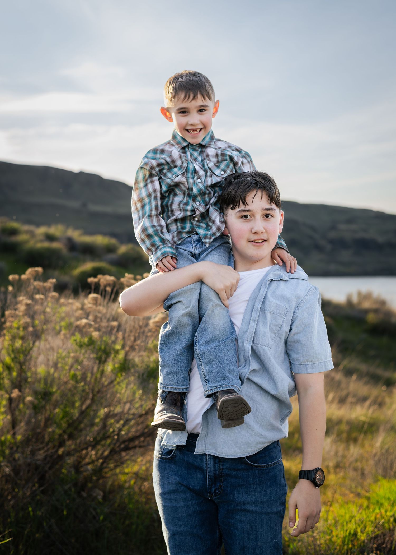 Two boys outdoors, older boy carrying younger sibling on shoulders, smiling near a lake with hills in background.