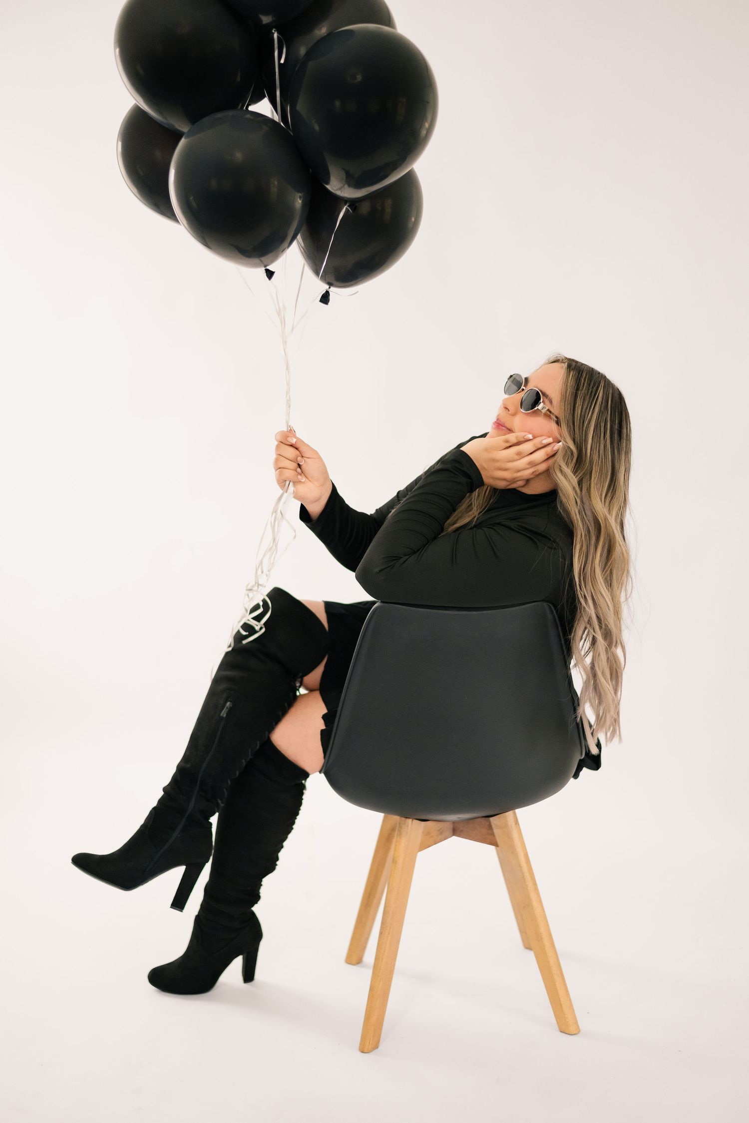 Stylish woman in black outfit sits on modern chair holding black balloons against white background.