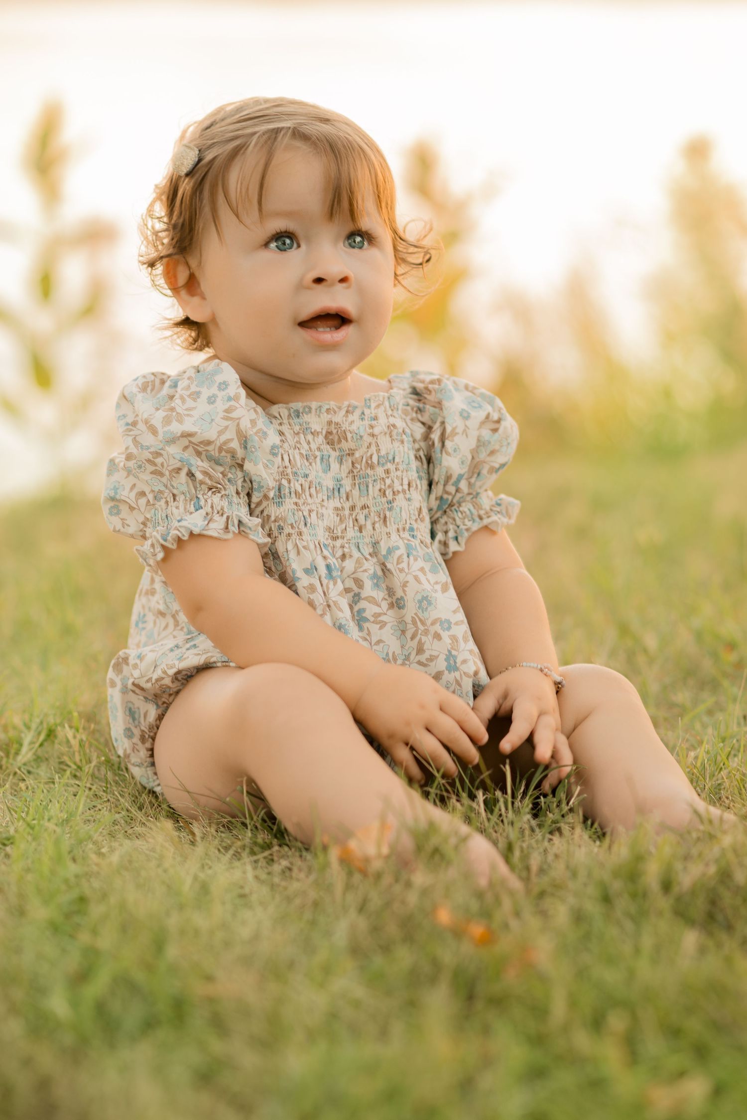 Sweet baby in floral dress sits on grassy hill during golden hour sunset.