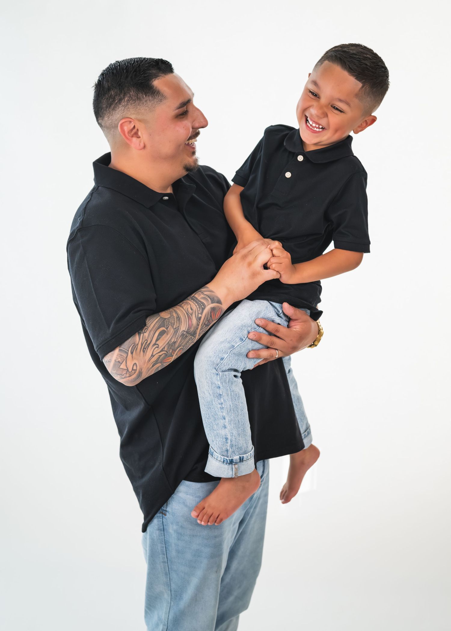 A tattooed father in a black polo shirt joyfully holds his laughing young son, both wearing matching outfits.