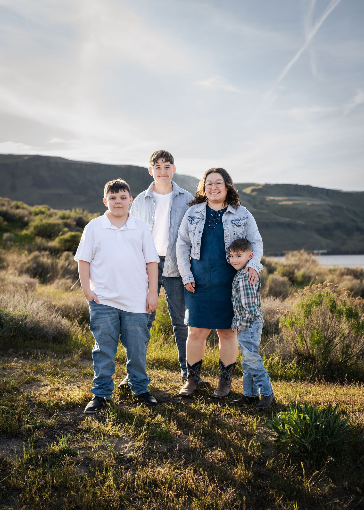 Family of four posing outdoors in a scenic landscape with hills and a lake in the background on a sunny day.
