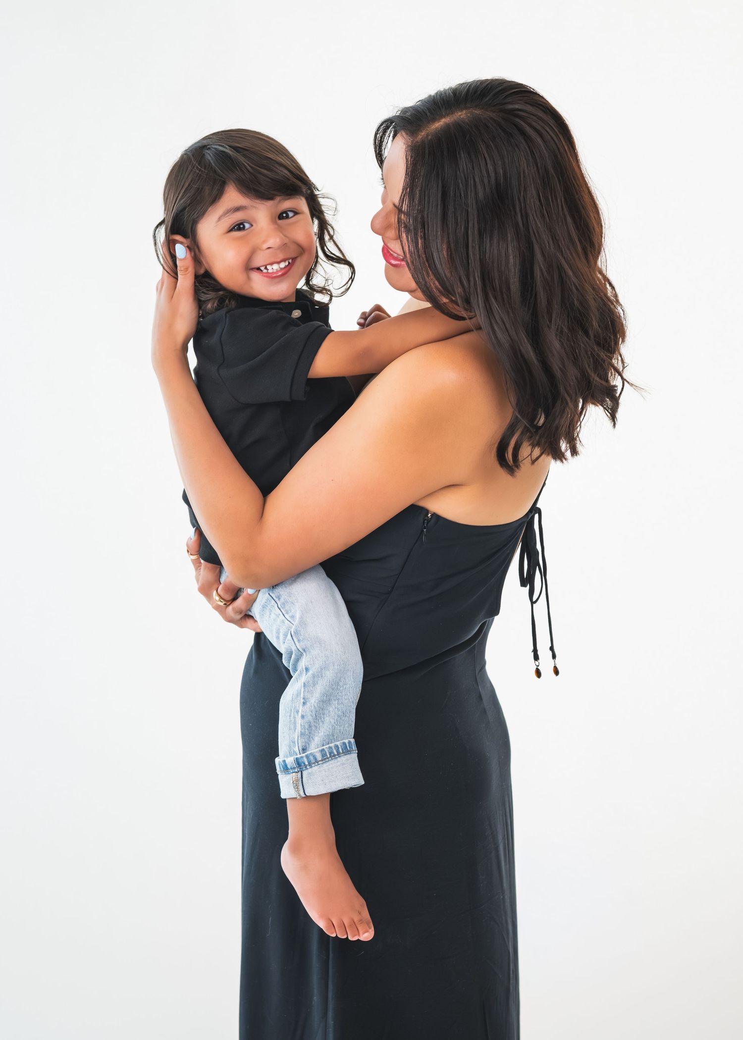 A woman in a black dress holds a smiling young child in a black shirt and jeans against a white background.