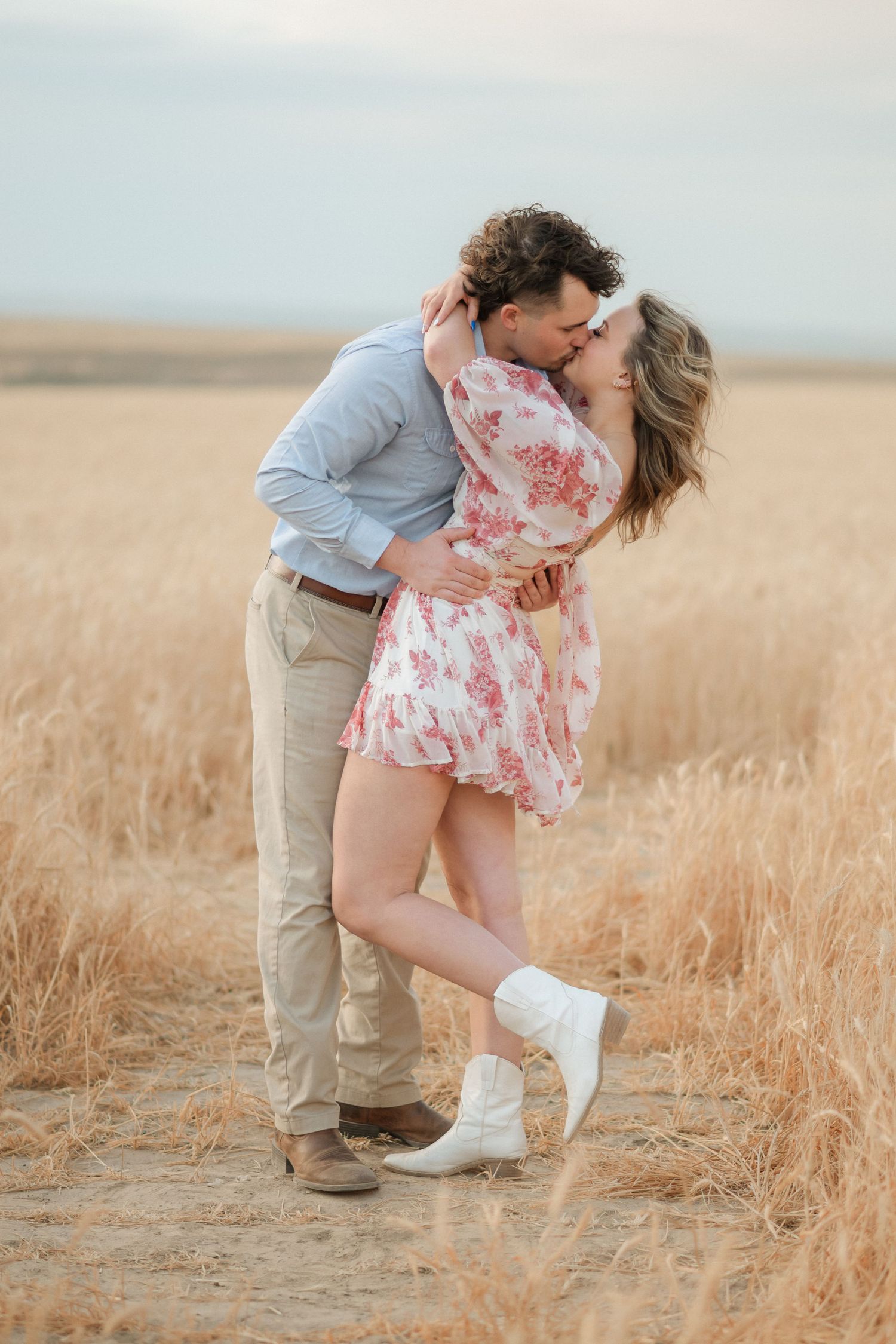 Romantic couple embraces and kisses in golden wheat field during sunset engagement photo session.