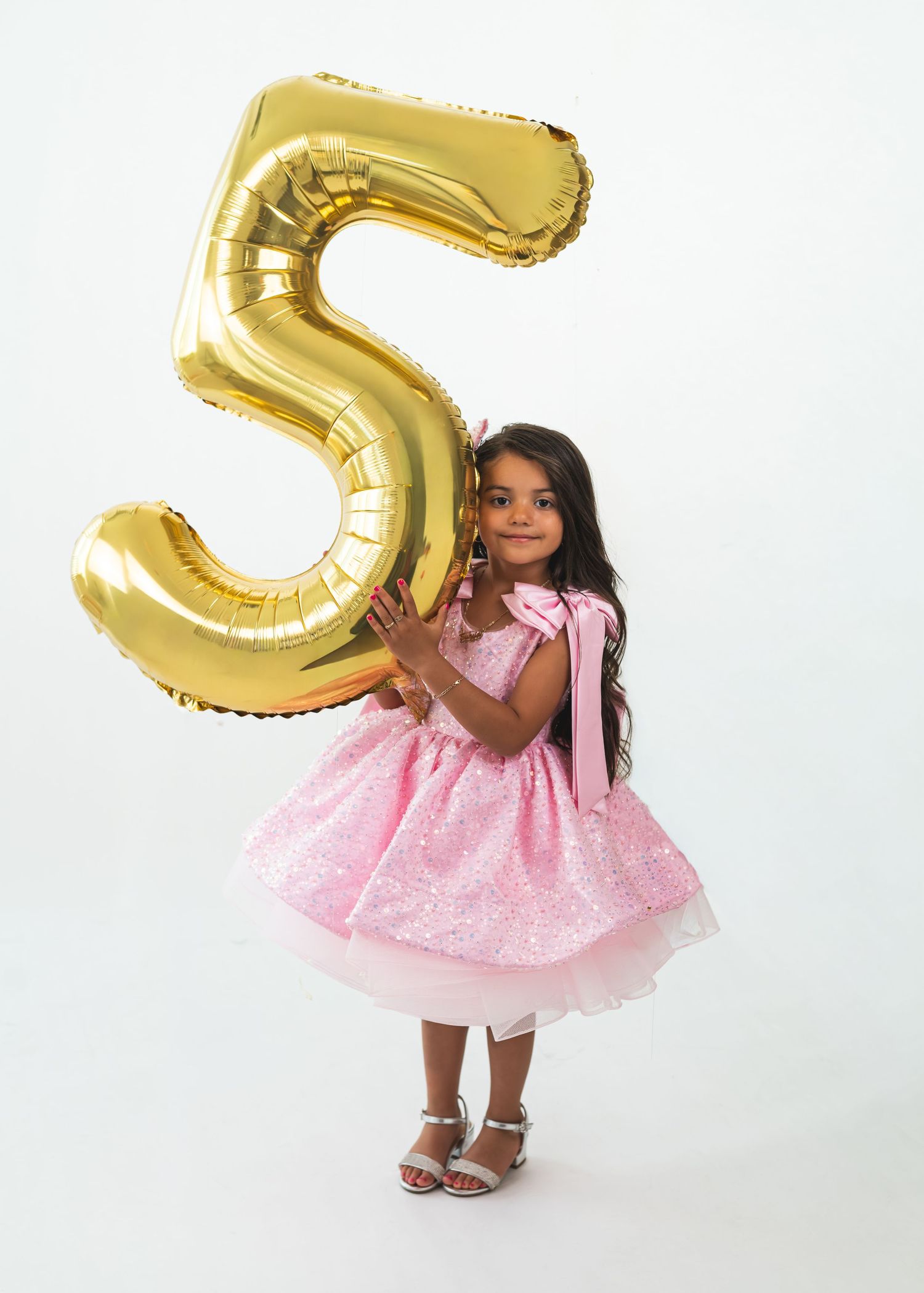 A young girl in a pink sequin dress holds a large gold number 5 balloon, celebrating her 5th birthday.