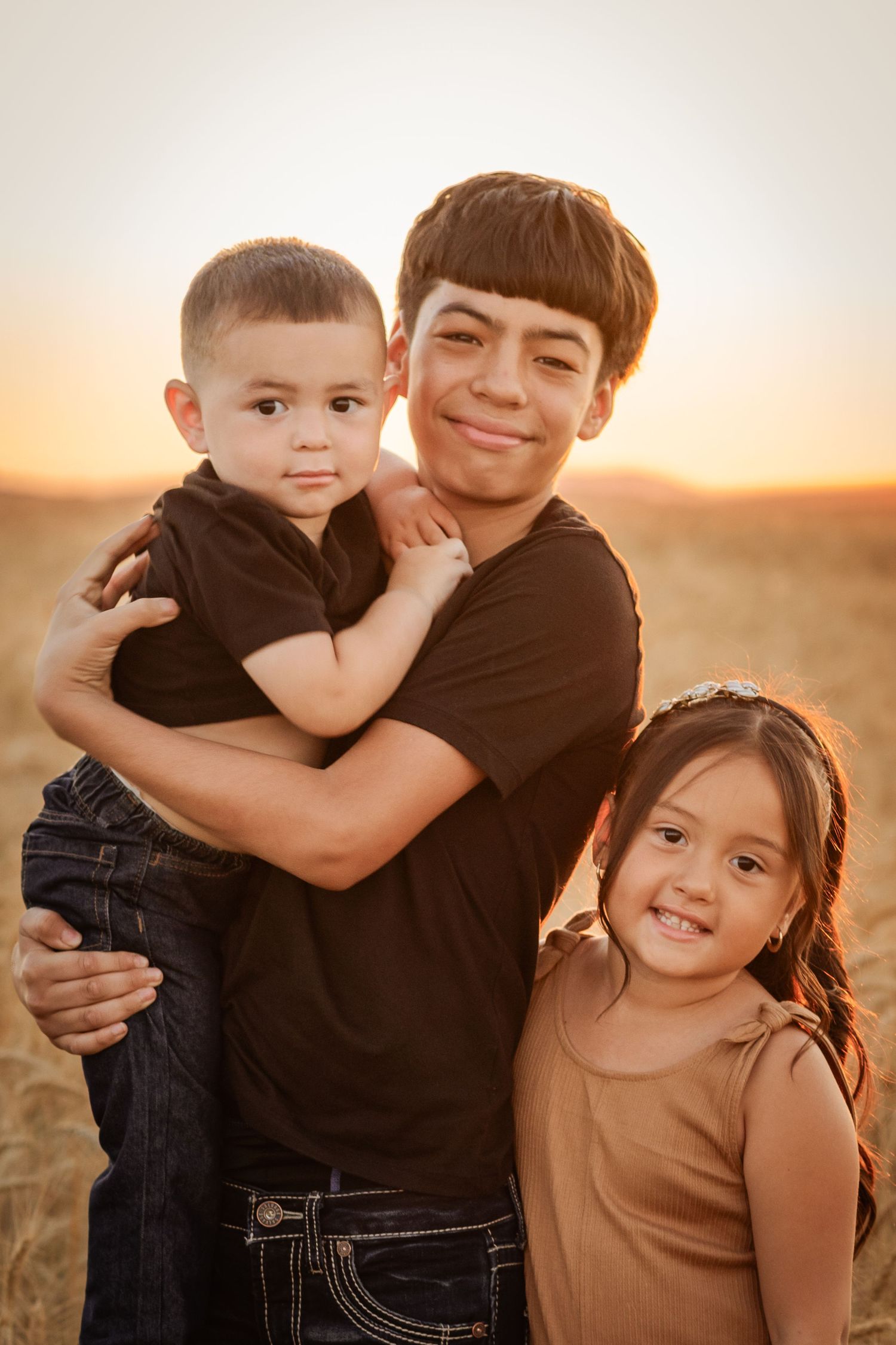 Three siblings pose at golden hour in a wheat field, older boy hugging toddler brother while smiling girl stands beside them.