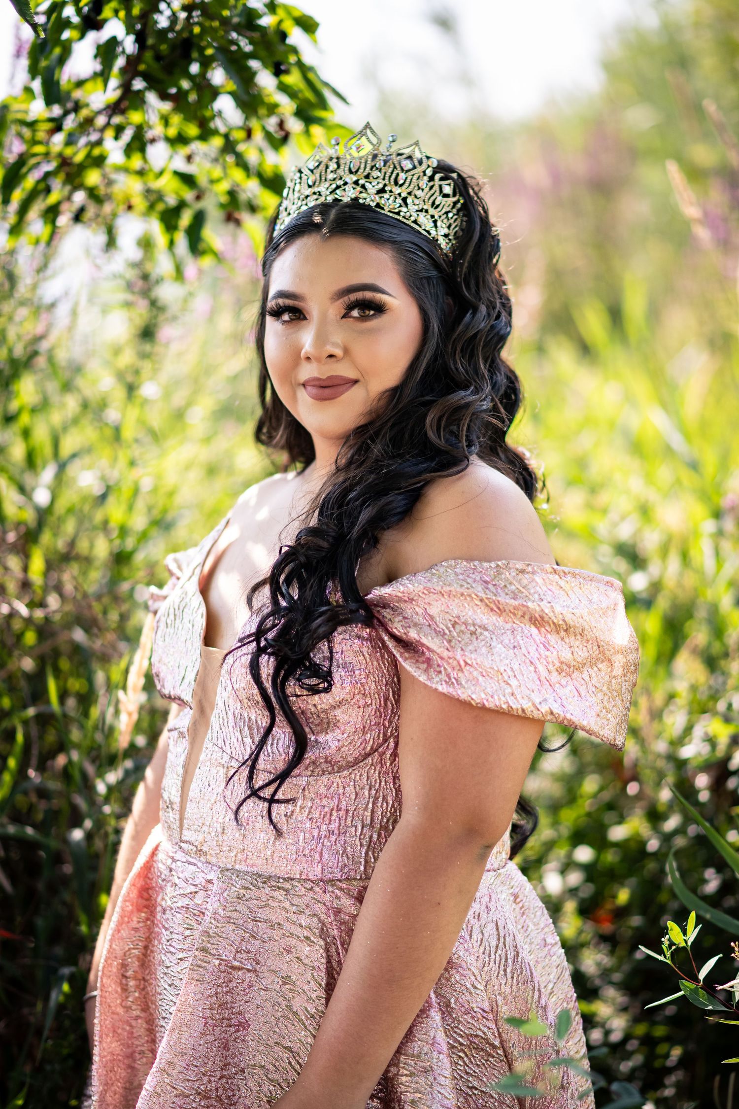 Young woman in pink off-shoulder gown and jeweled crown poses gracefully outdoors among lush greenery.