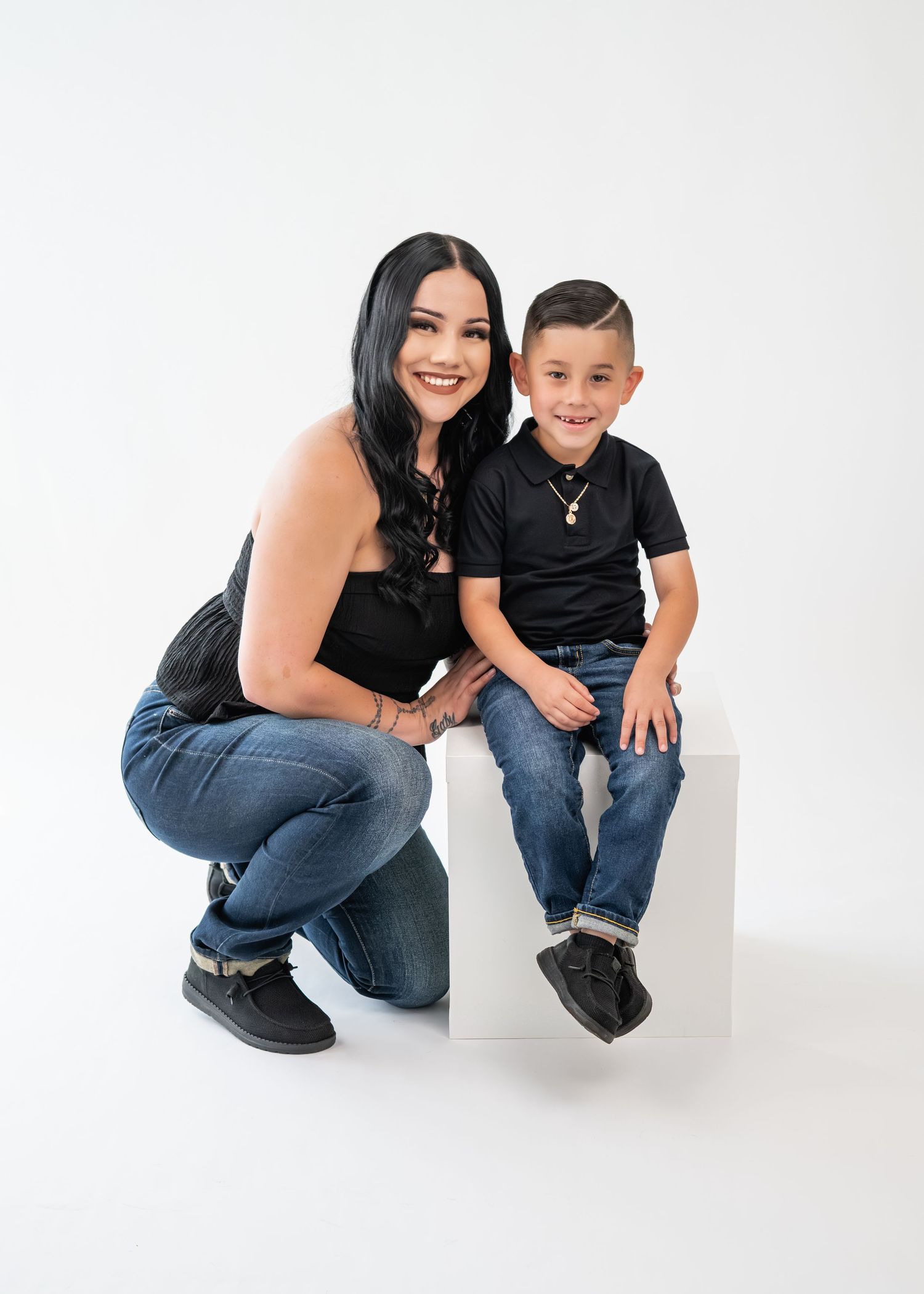 A woman and young boy in matching black outfits and jeans pose together on a white cube against a white background.