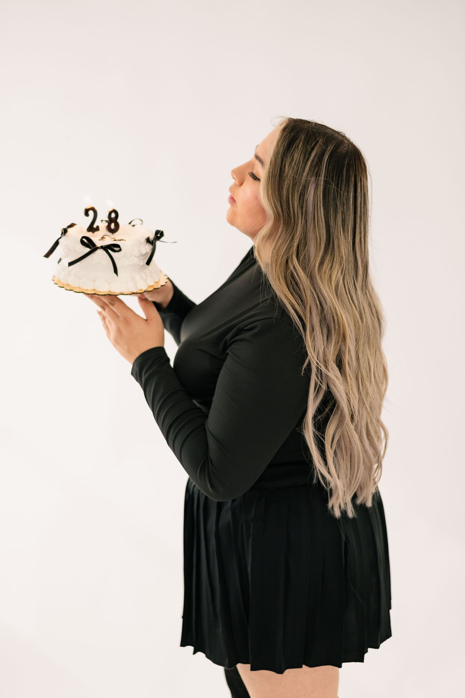 Woman in black dress holding a white birthday cake with number 28 on top against white background.
