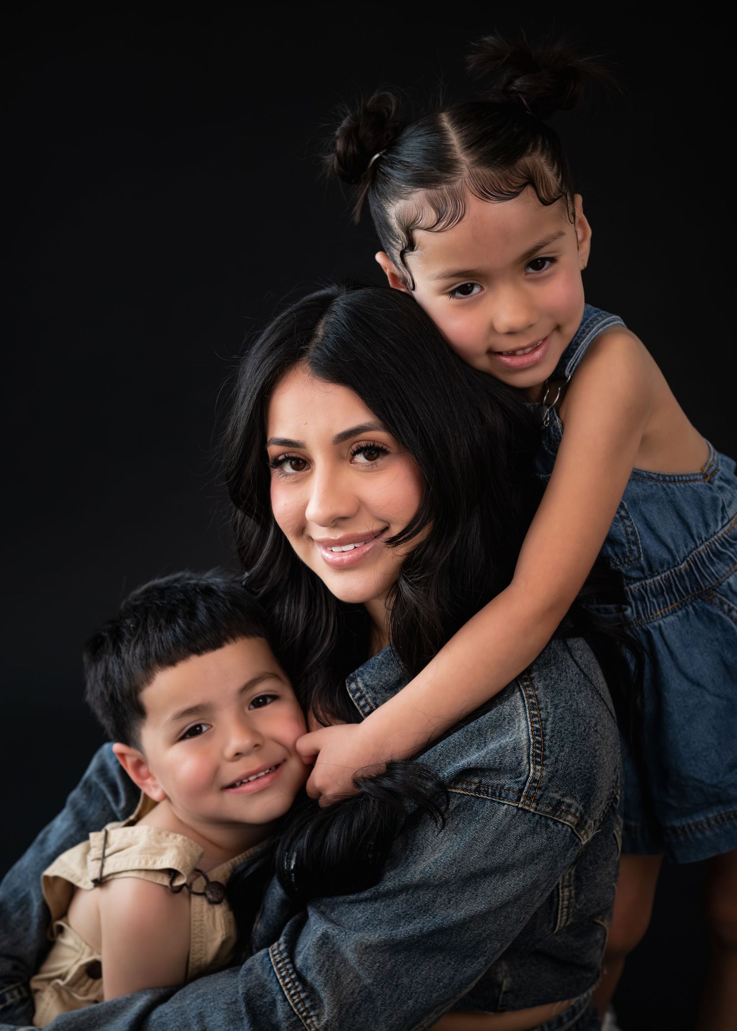 A smiling mother poses with her two young children against a dark background in a warm family portrait.