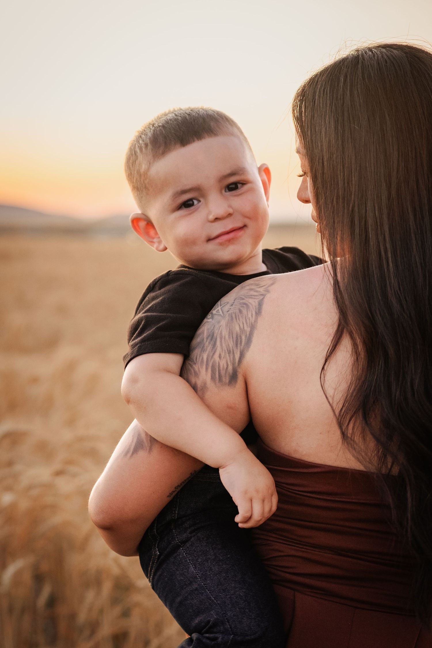 A smiling toddler boy in a black shirt being held by a tattooed woman in a golden wheat field at sunset.