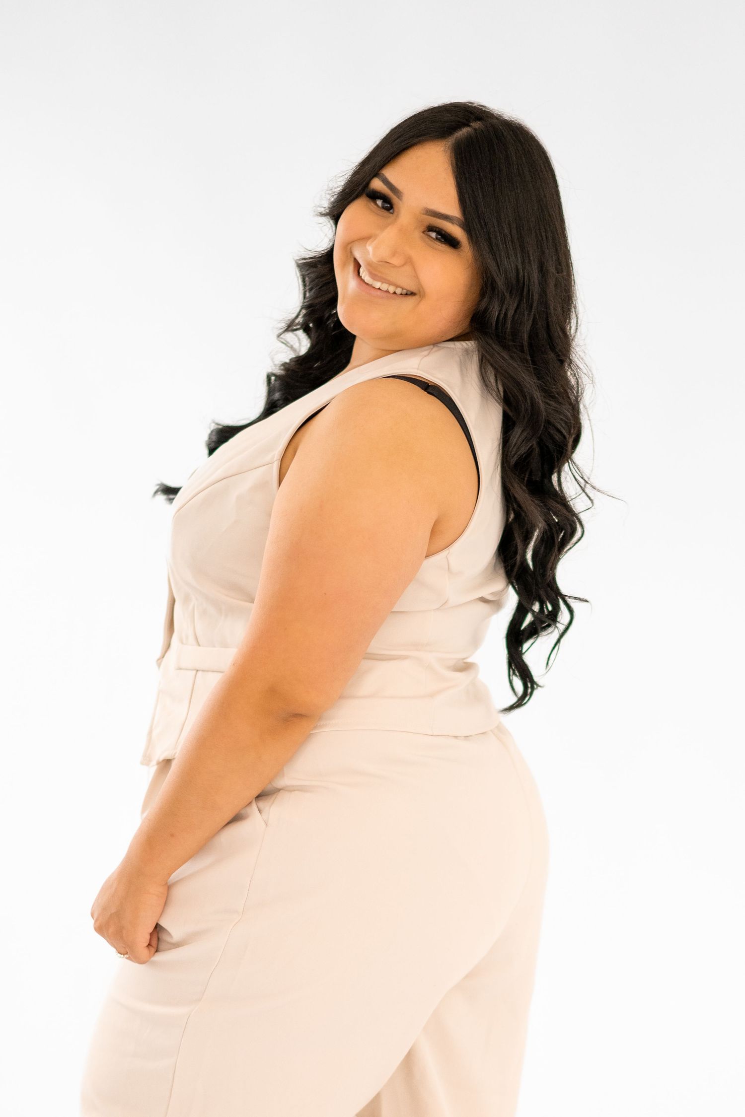 Smiling woman in cream-colored sleeveless outfit poses against white background with long dark wavy hair.