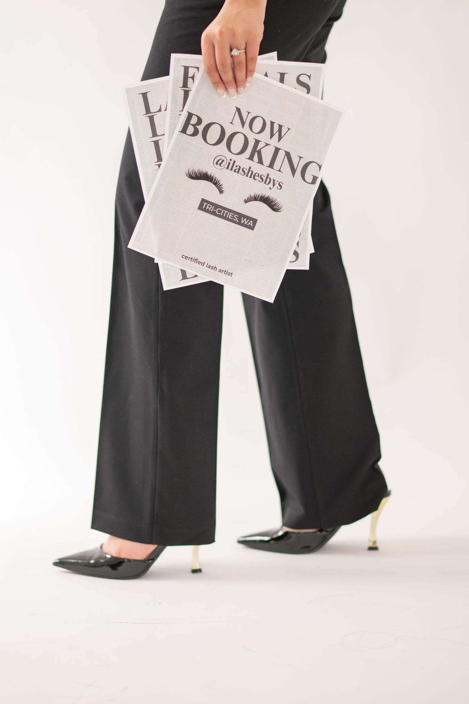 Person wearing black wide-leg pants and pointed heels holds papers with 'Now Booking' text against white background.