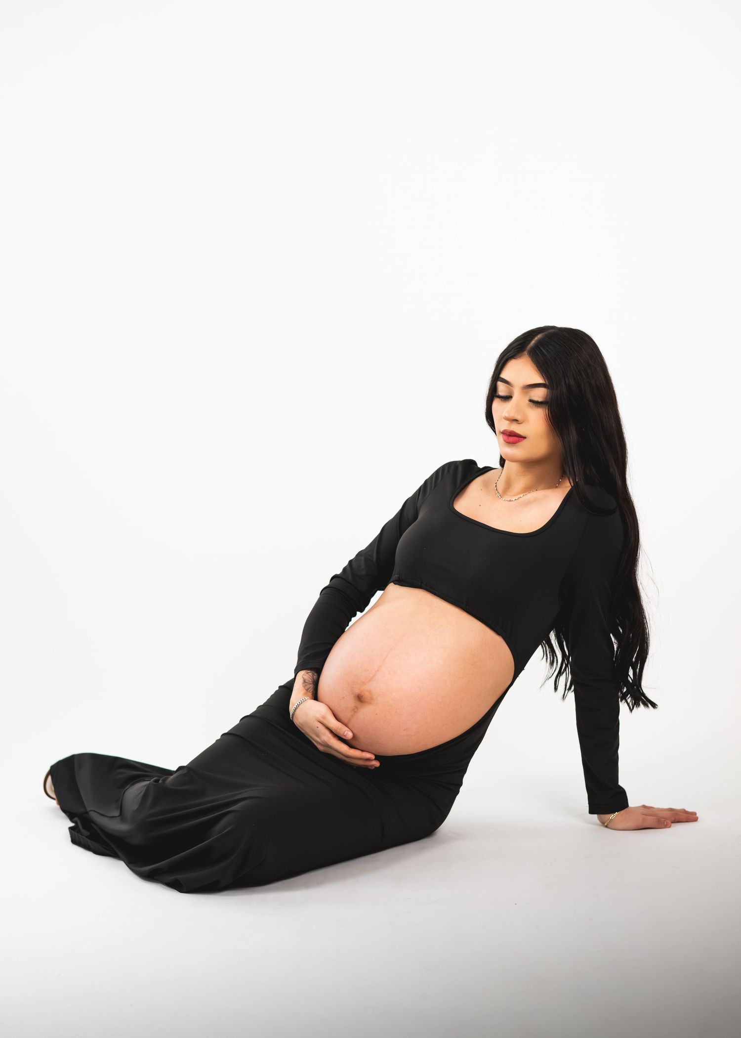 Pregnant woman in black outfit sitting gracefully on white background, cradling her baby bump with long dark hair.