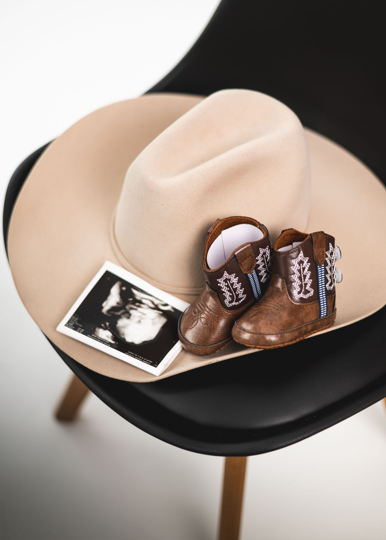 Baby cowboy boots and ultrasound photo on a tan cowboy hat atop a black modern chair, pregnancy announcement.