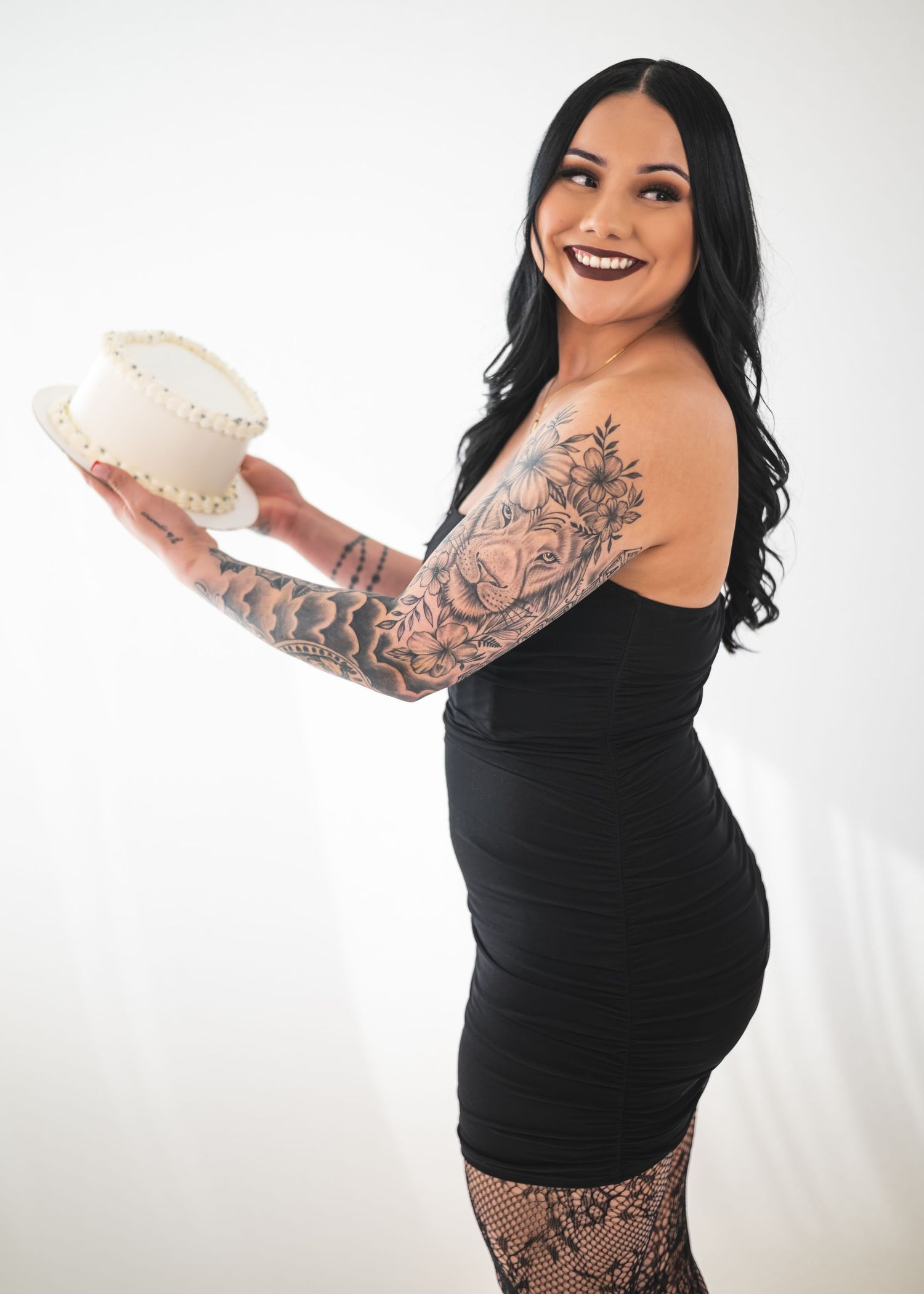 Tattooed woman in black dress holding a white cake, smiling over shoulder against white background.