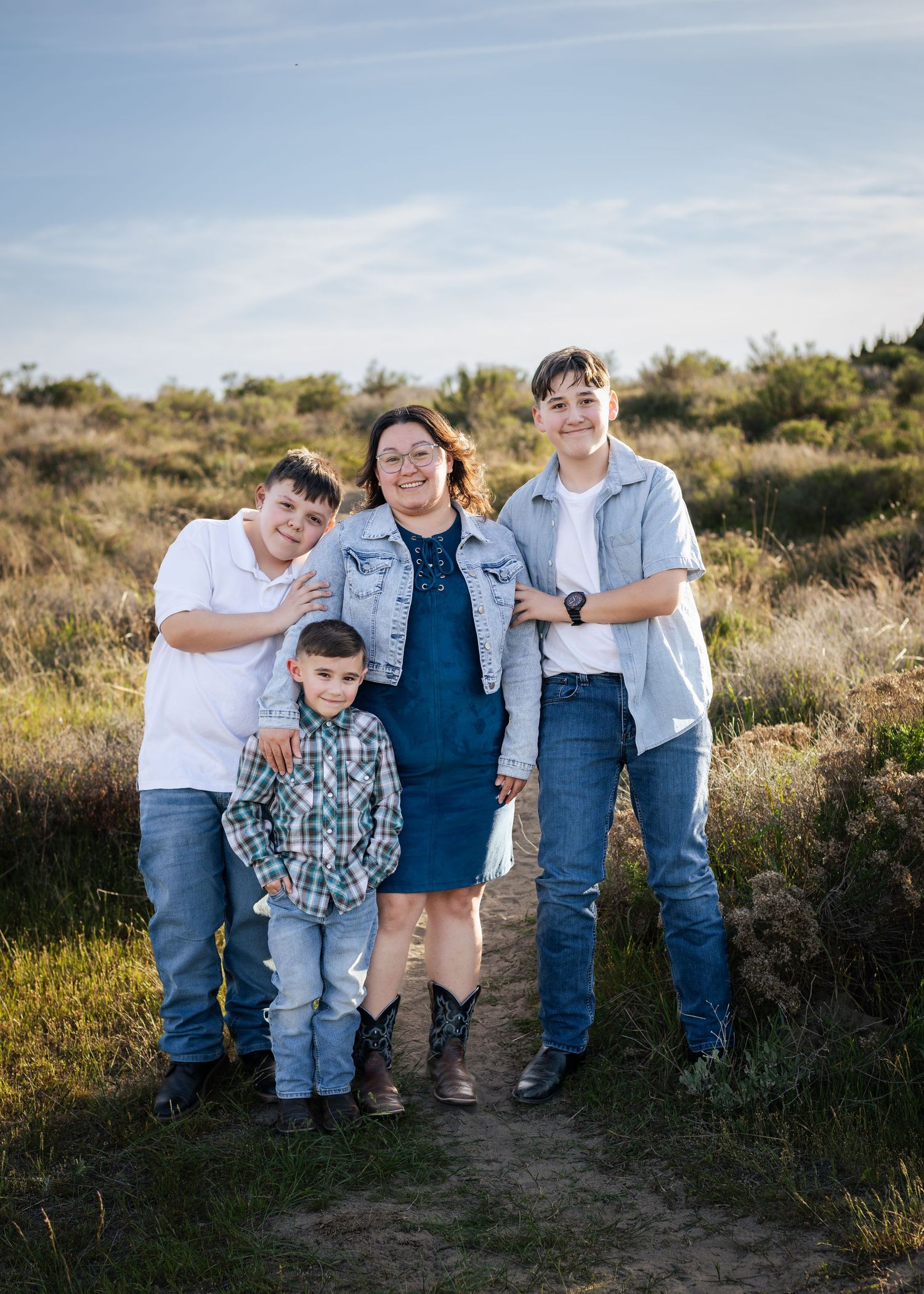 A woman and three boys pose together on a nature trail, dressed in denim and western wear, smiling outdoors.