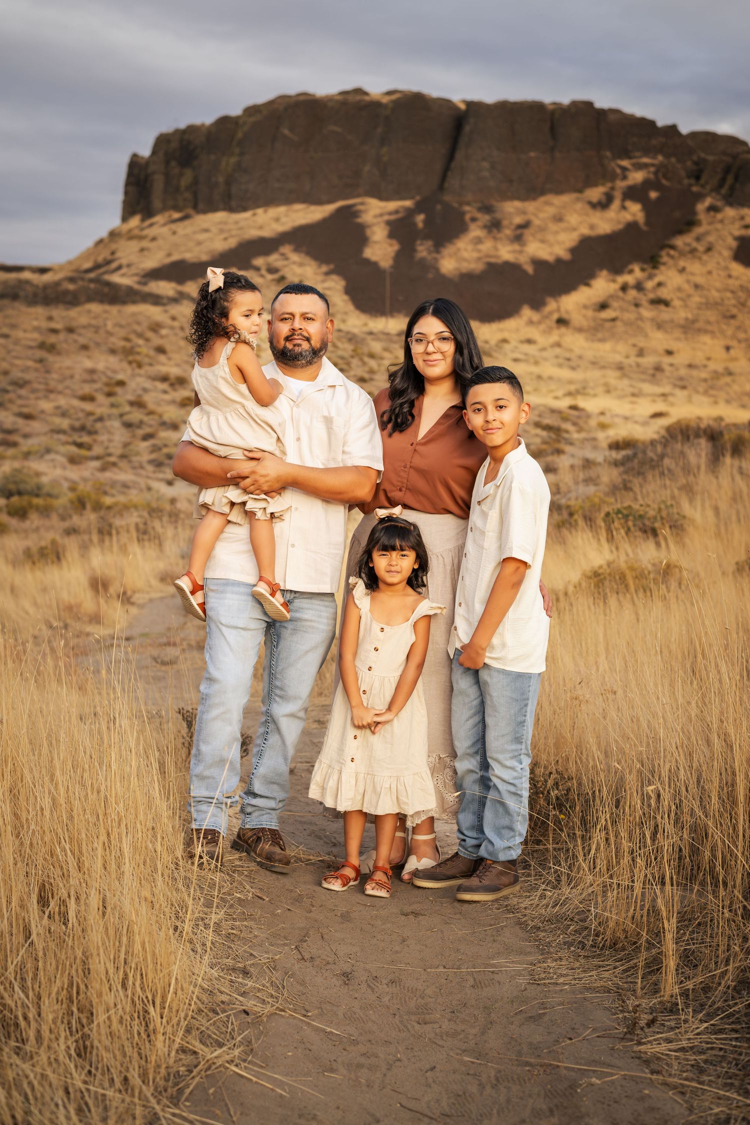 A family wearing coordinated cream and denim outfits poses together in a desert landscape with golden grass at sunset.