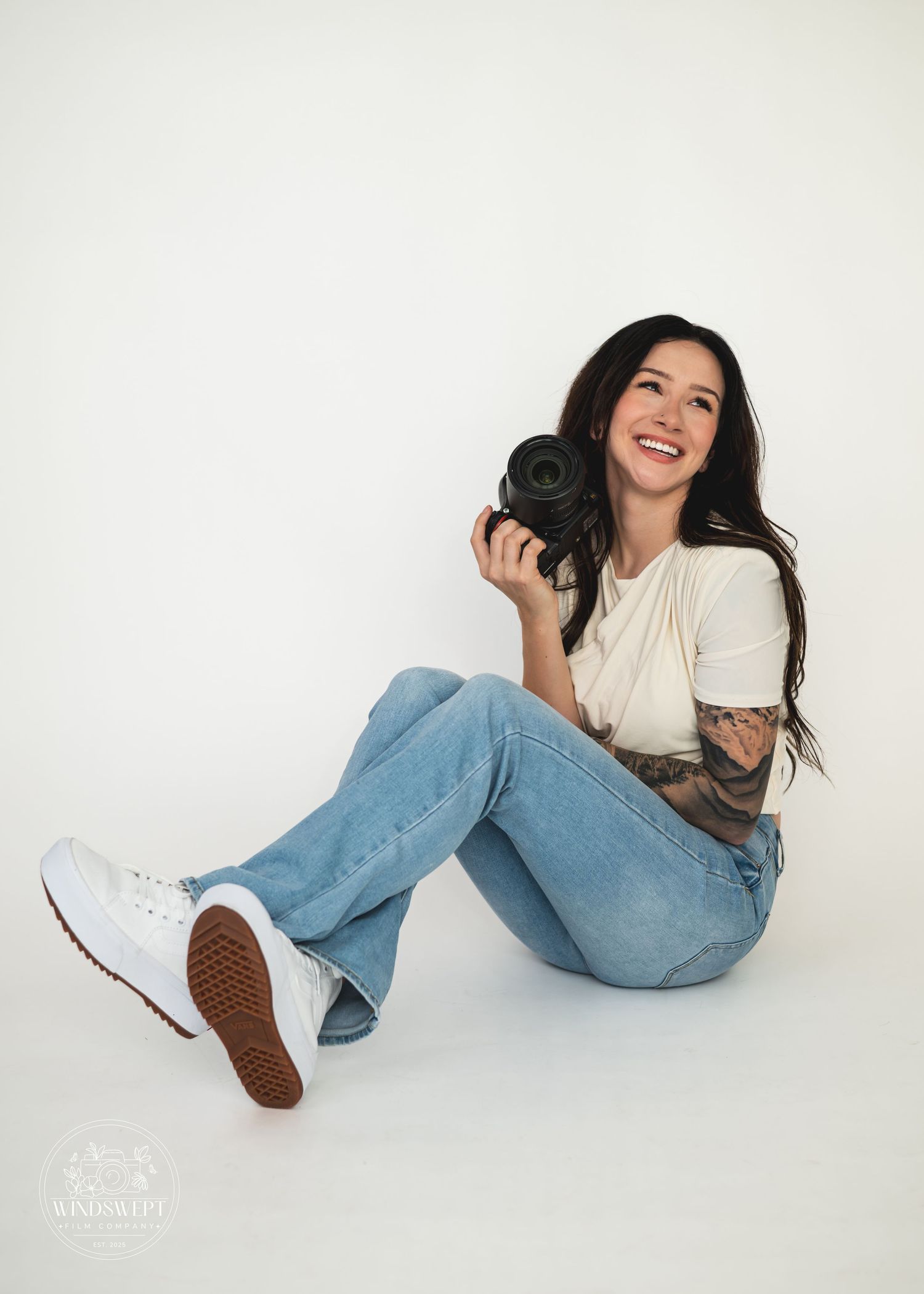 Photographer in white sweater and jeans poses playfully with camera against white backdrop in creative studio shots.