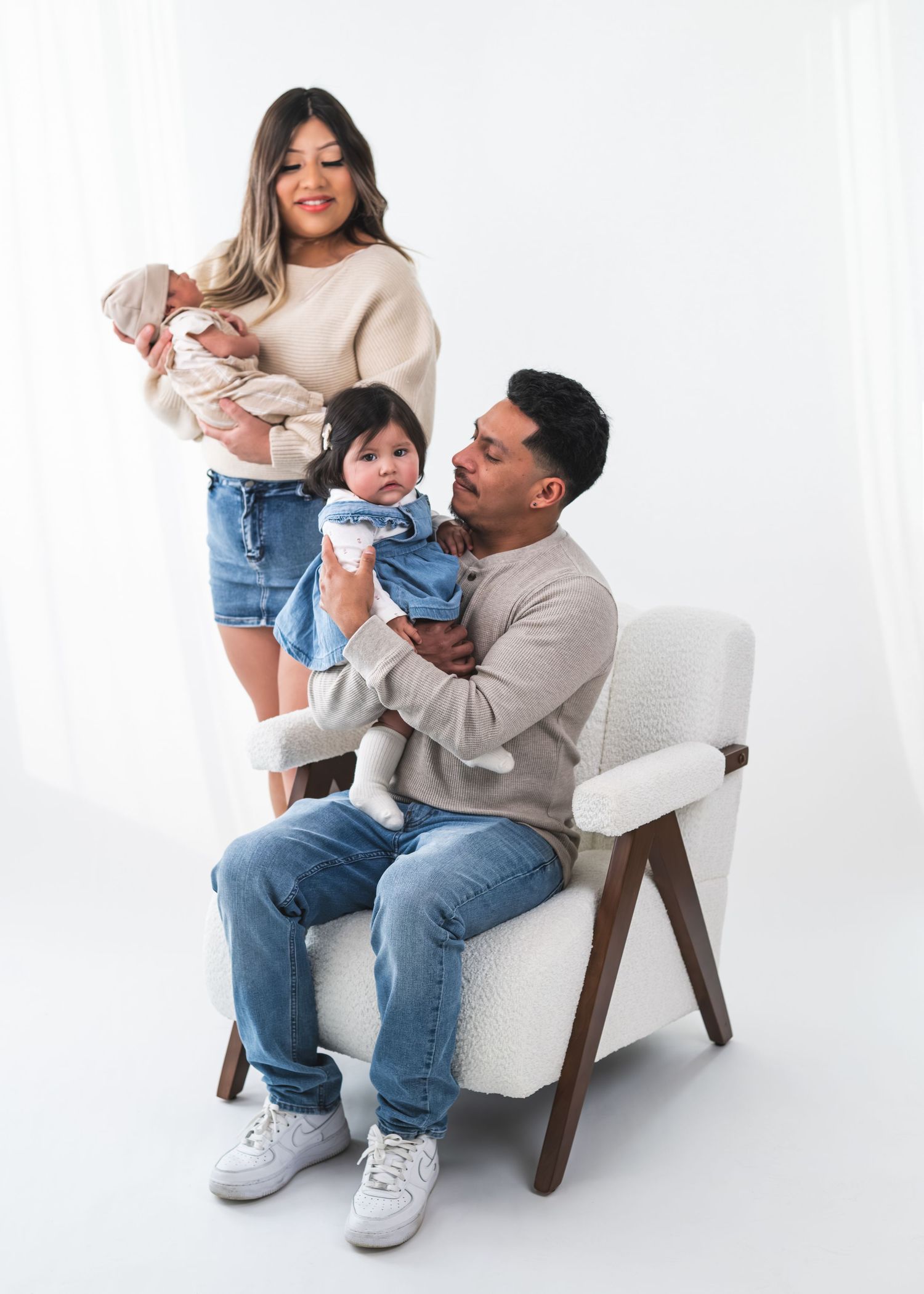 A young family poses in a studio, mom holding a newborn, dad seated cradling a baby girl, all in casual neutral tones.