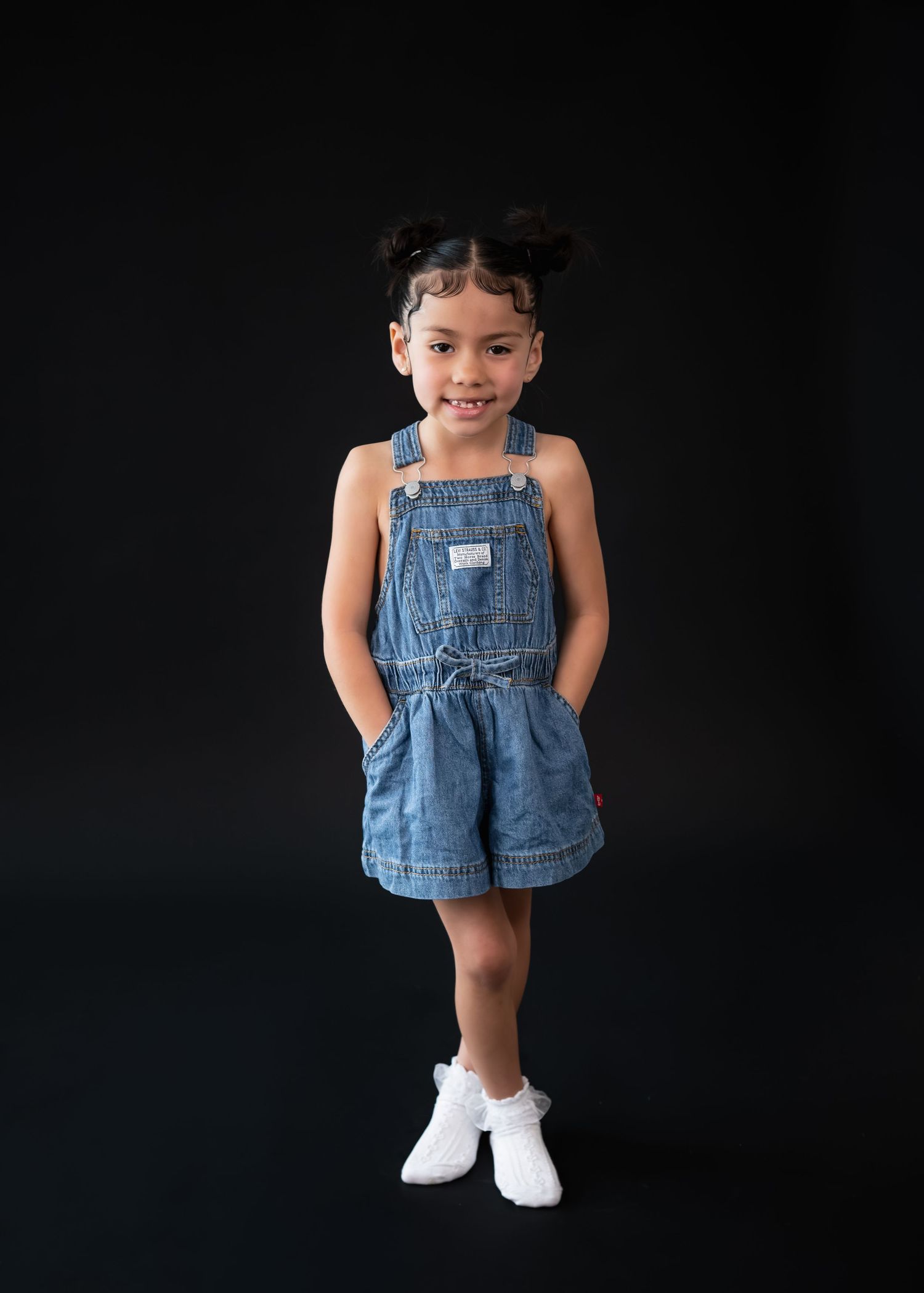 A smiling young girl in denim overalls with pigtails poses confidently against a black background.