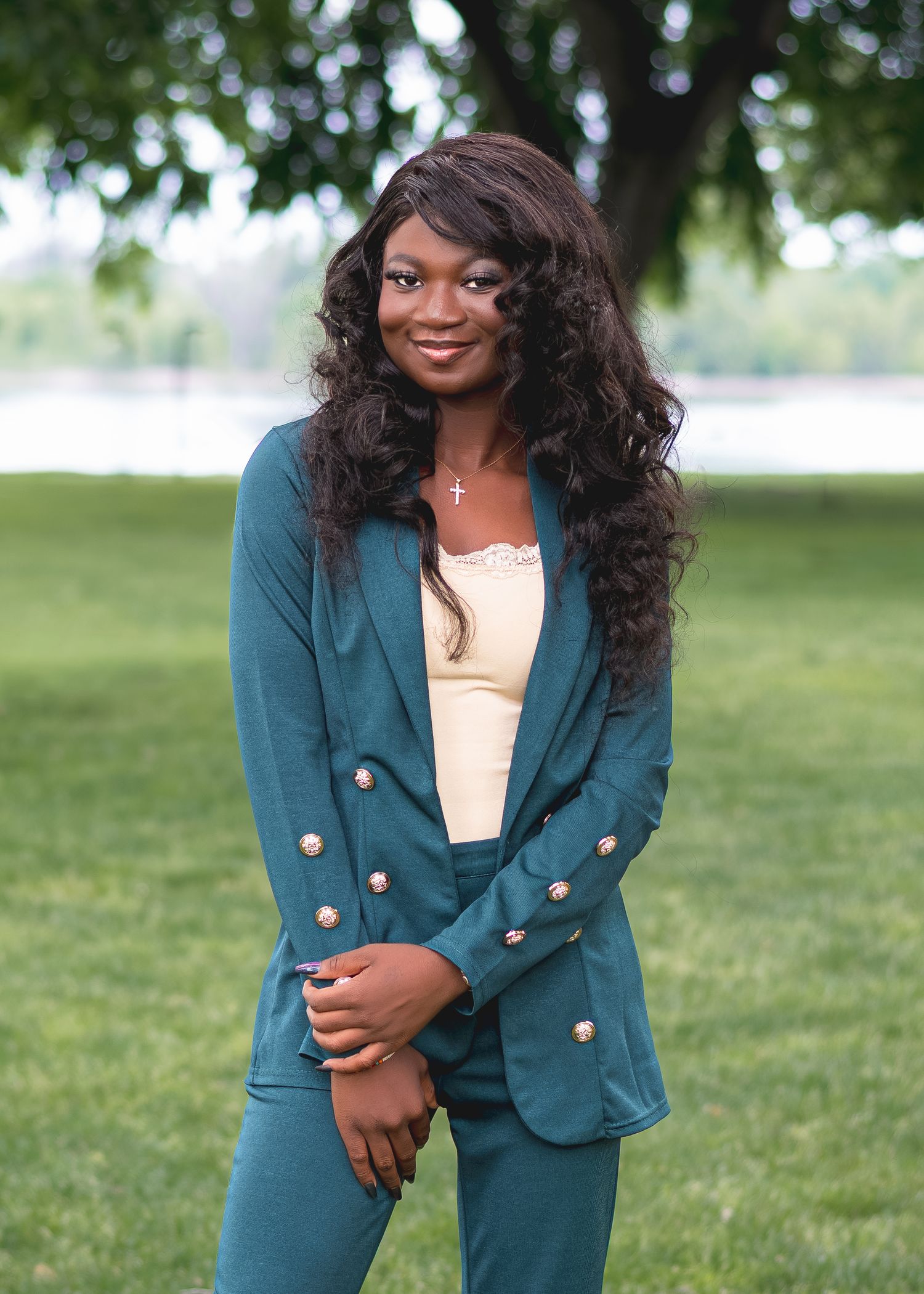 Young woman in teal double-breasted suit posing outdoors in a park with green trees and water in background.