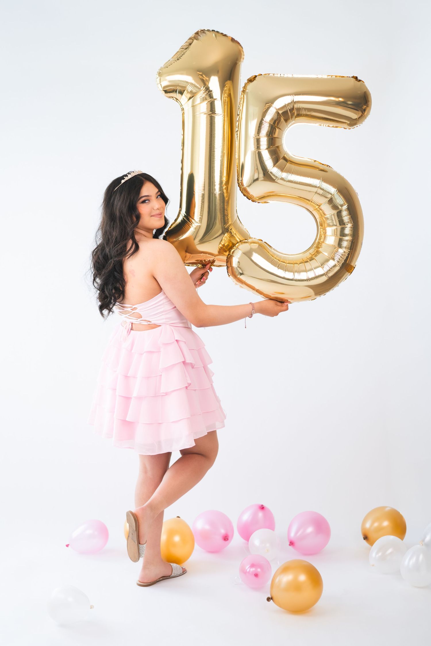 Young woman in pink ruffled dress holding gold number 15 balloons, celebrating quinceañera with pink and gold balloons on floor.
