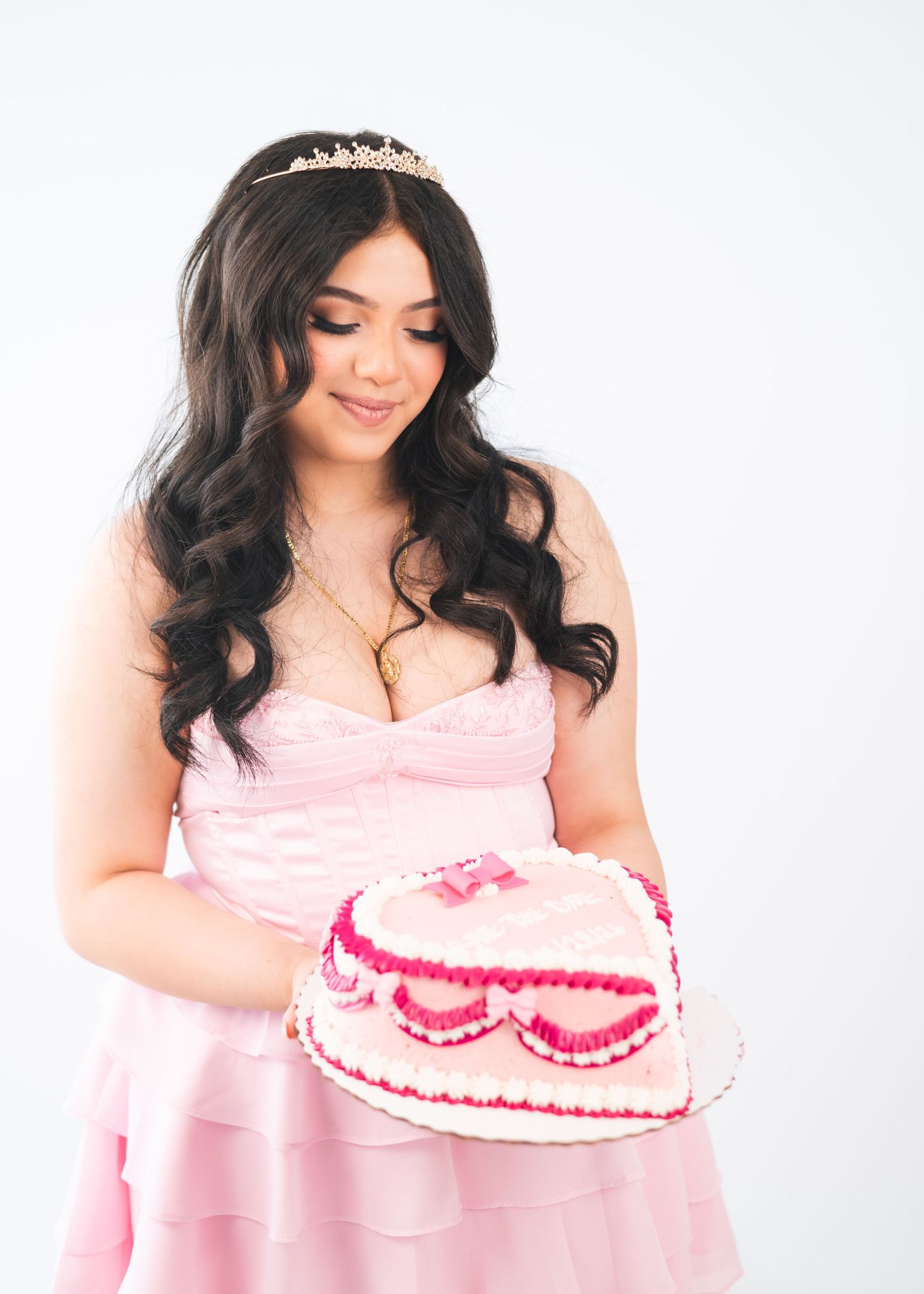 Young woman in pink gown and tiara holding a heart-shaped pink birthday cake against white background.
