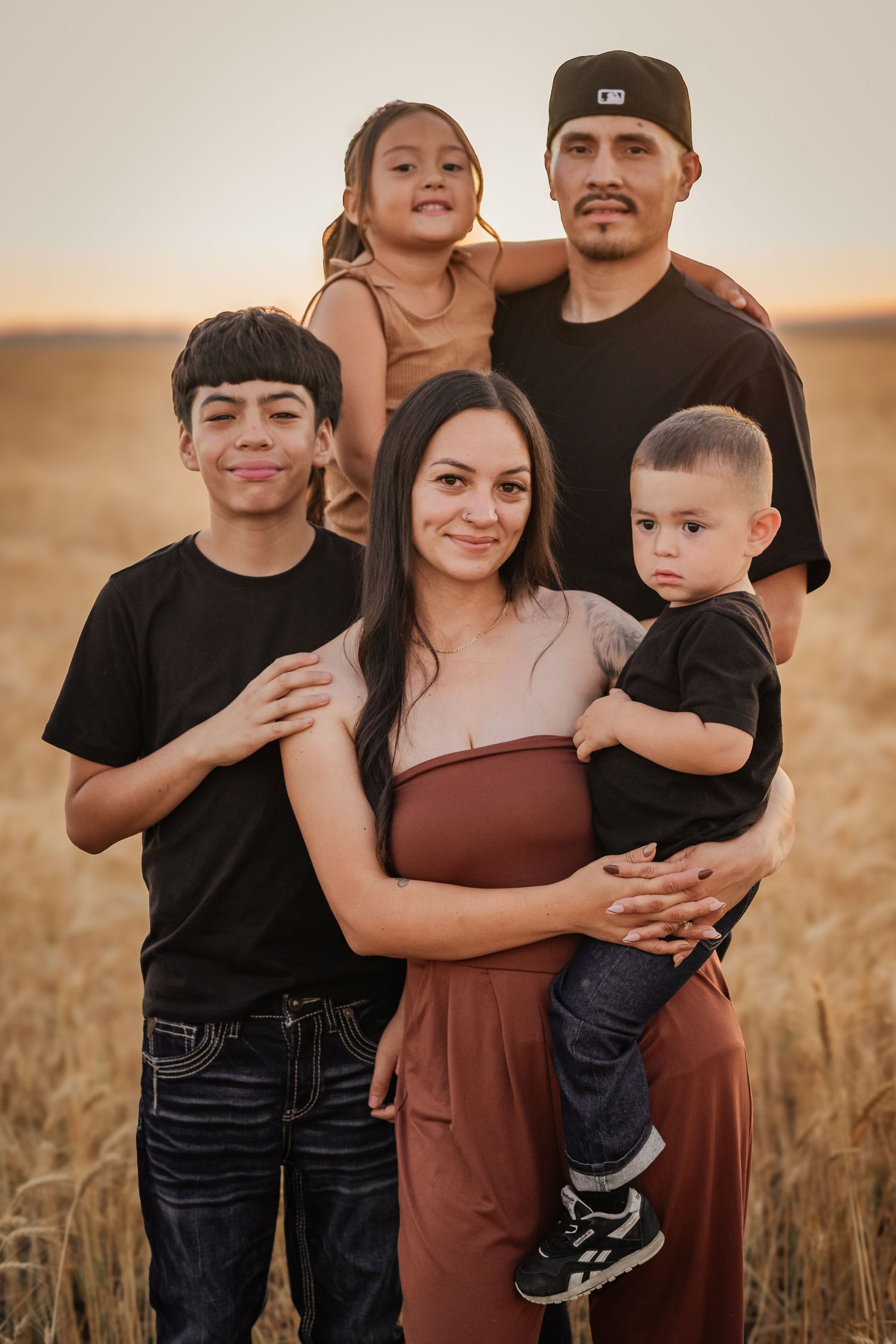 Family of five posing together in a golden wheat field at sunset, smiling warmly for a portrait photo.