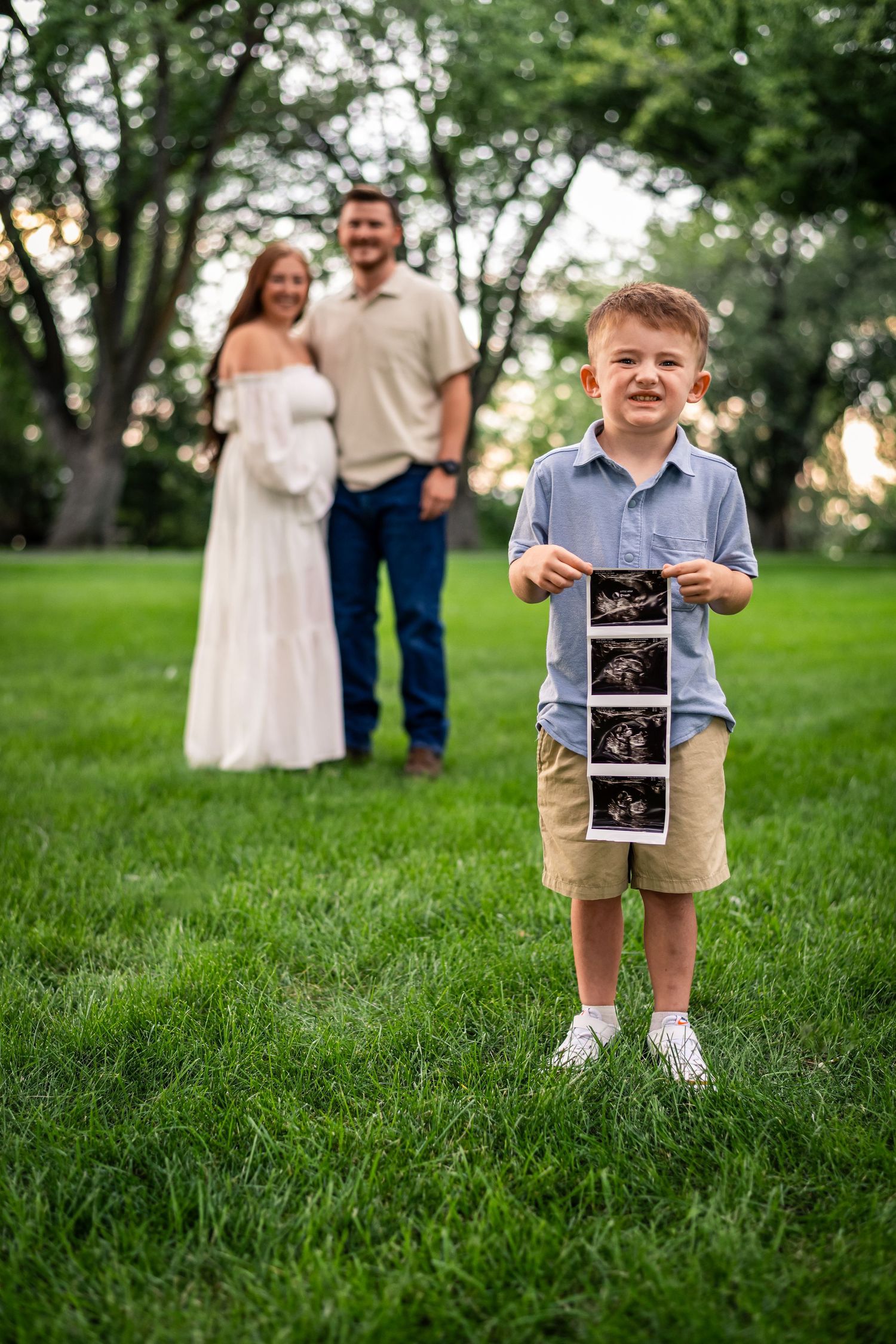 Young child stands in grassy field holding ultrasound photos while couple stands in background during sunset.
