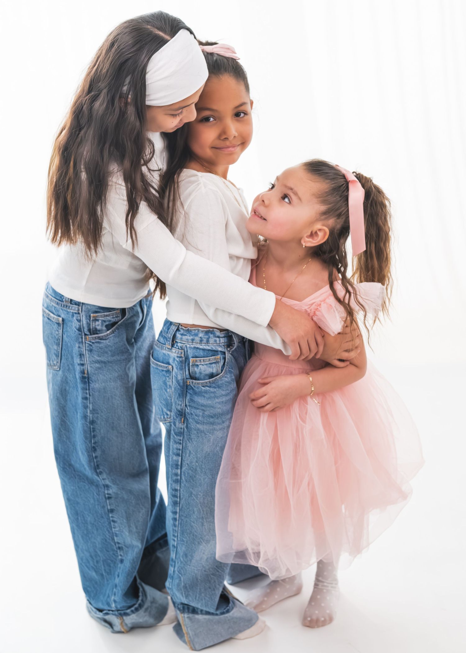 Three young girls hugging and smiling together, wearing jeans and a pink tutu dress, against a white background.