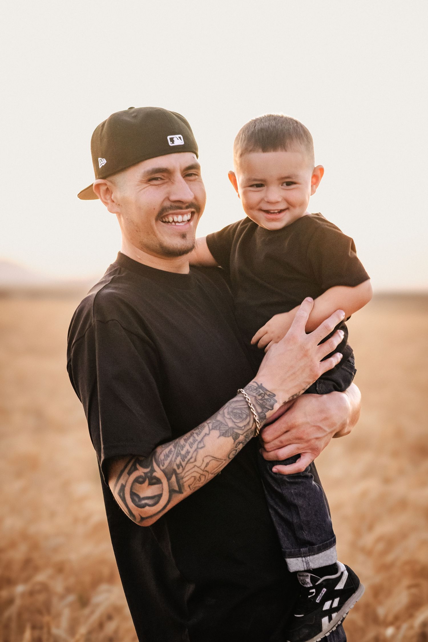 Smiling tattooed man in black tee and cap holding happy young boy in wheat field at golden hour.