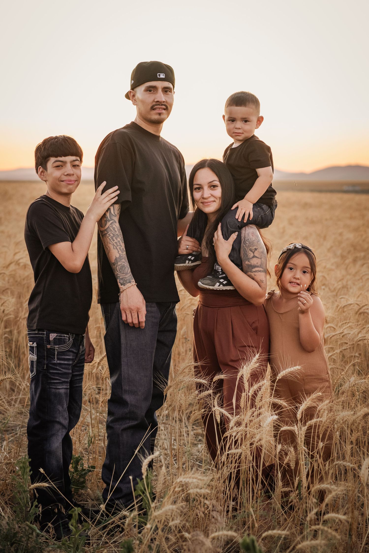 Family of five posing in golden wheat field at sunset, wearing black and brown outfits, with mountains in background.