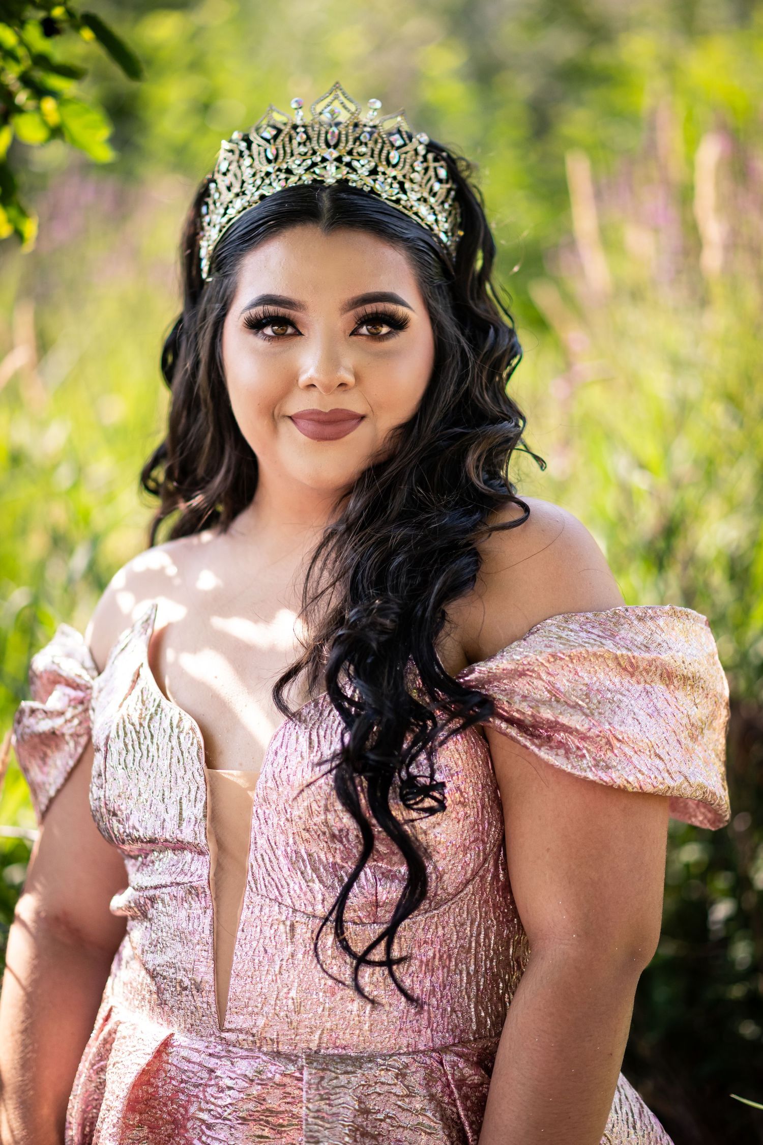 A person wearing a pink gown and crystal tiara poses outdoors in natural sunlight against a blurred green background.