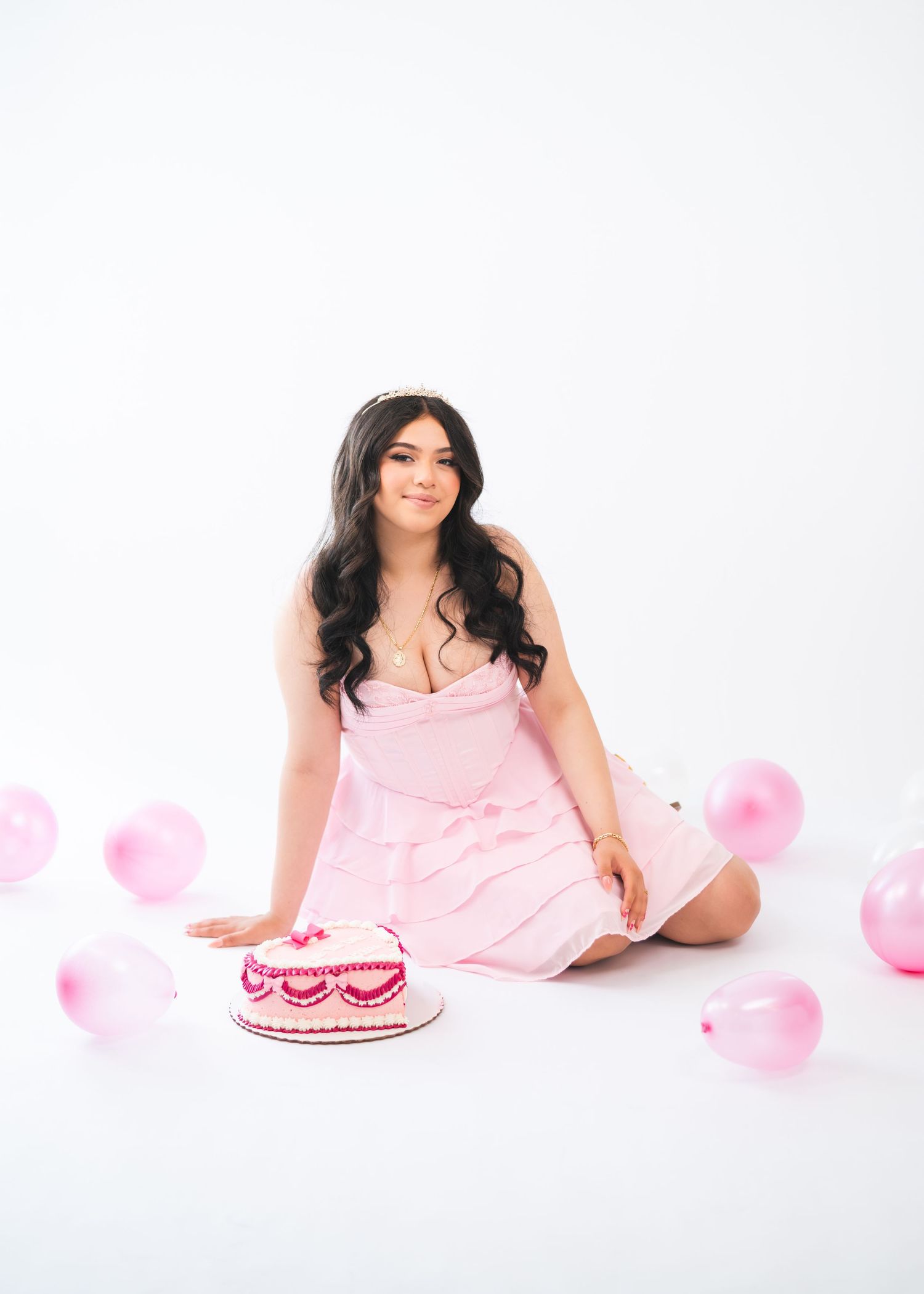 Woman in pink dress sitting on white floor with pink birthday cake and balloons around her.